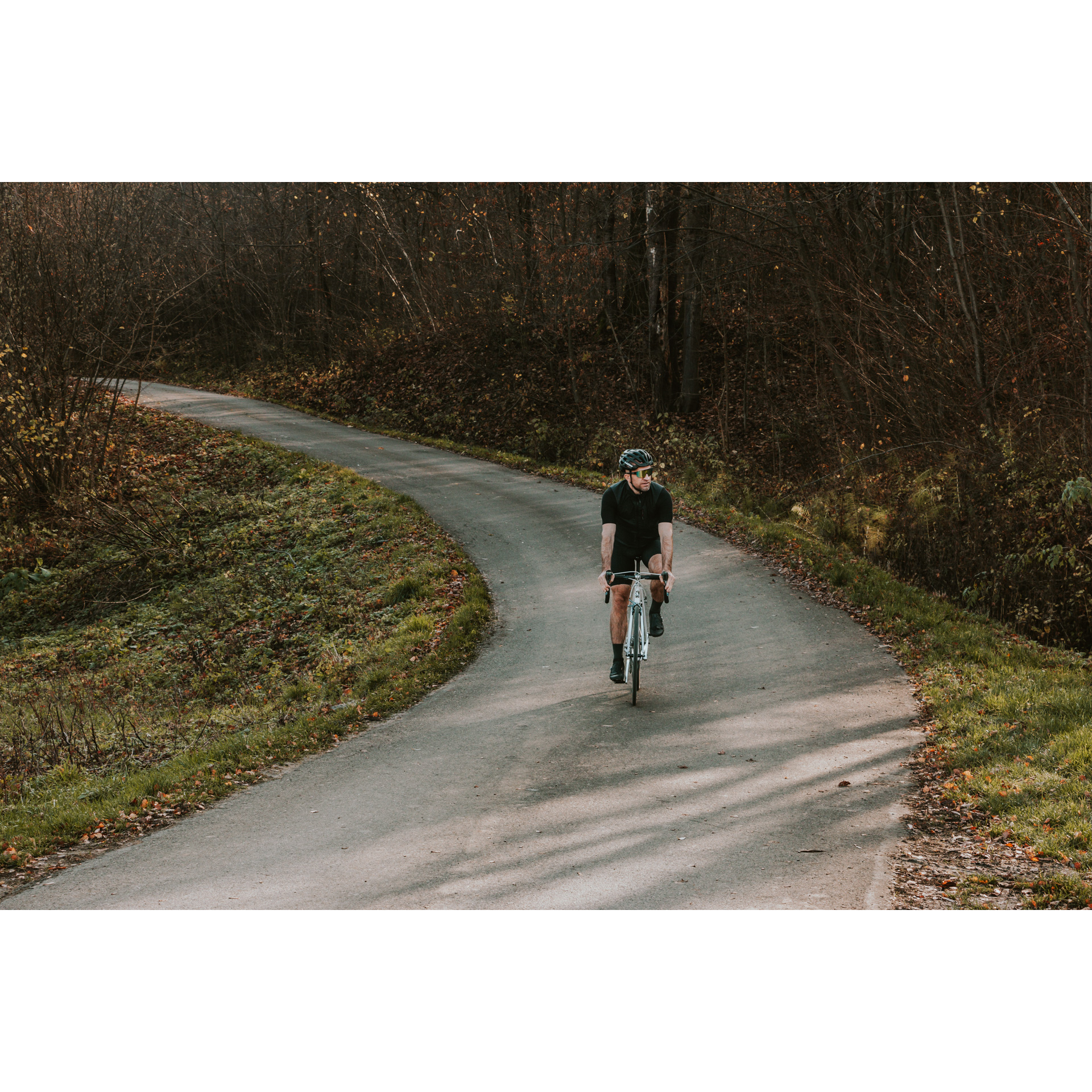 Kolarz w czarnym stroju, kasku i ciemnych okularach, jadący na rowerze asfaltową drogą biegnącą wśród drzew