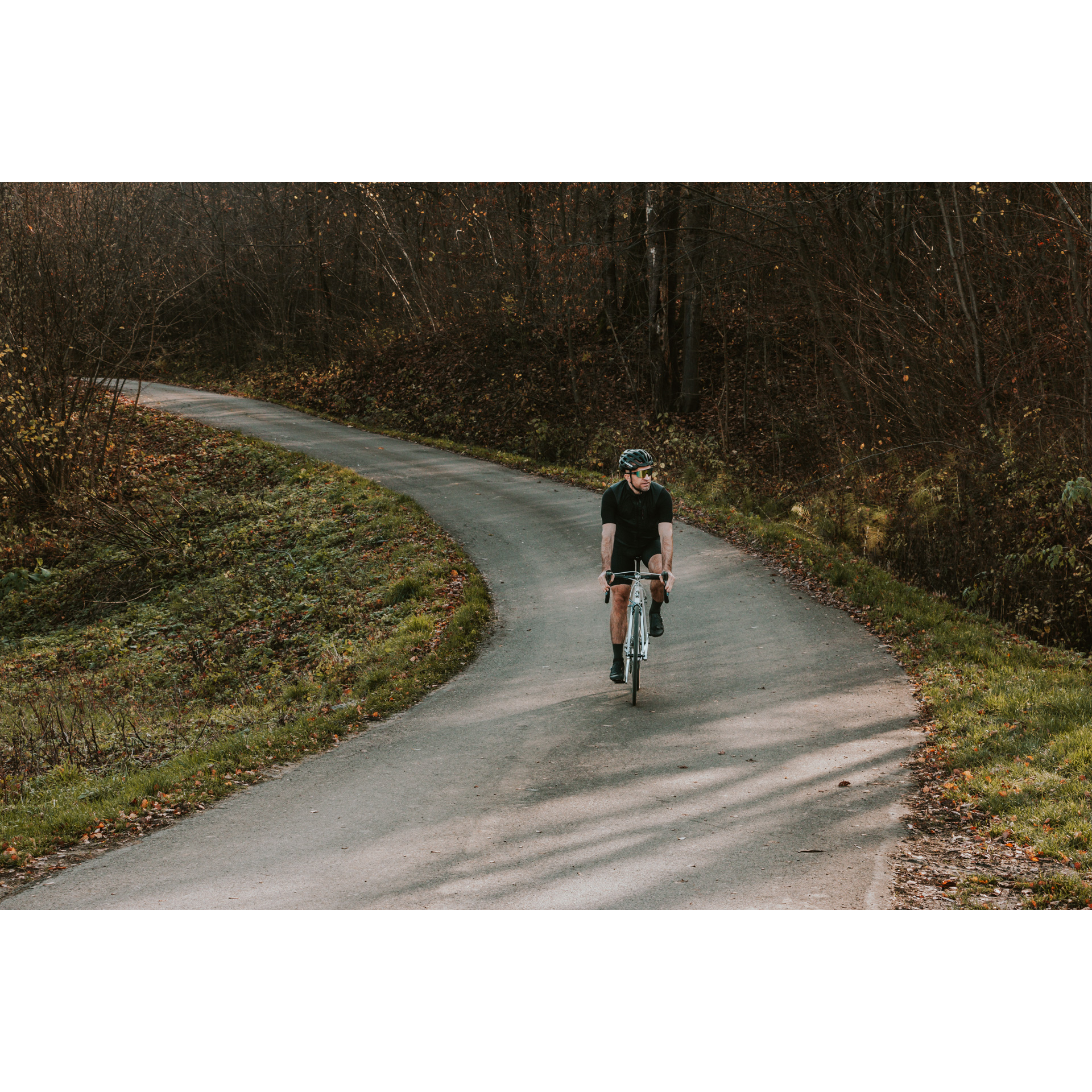 A cyclist in a black outfit, helmet and dark glasses, cycling along an asphalt road running among the trees