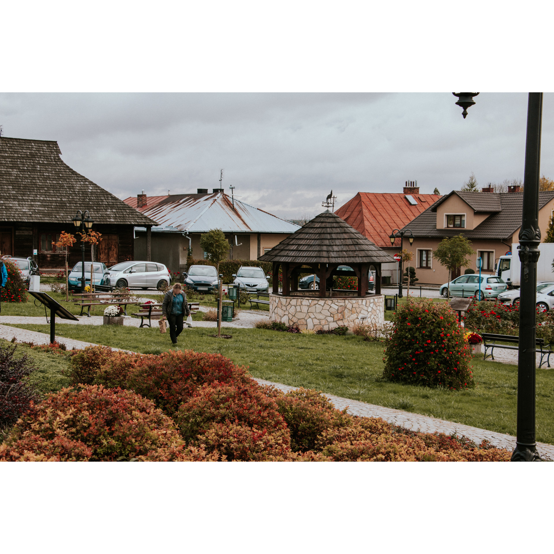 A brick, round well with a roof on a green pass, in the middle of the town, parked cars and buildings in the background, colorful bushes and a path in the foreground