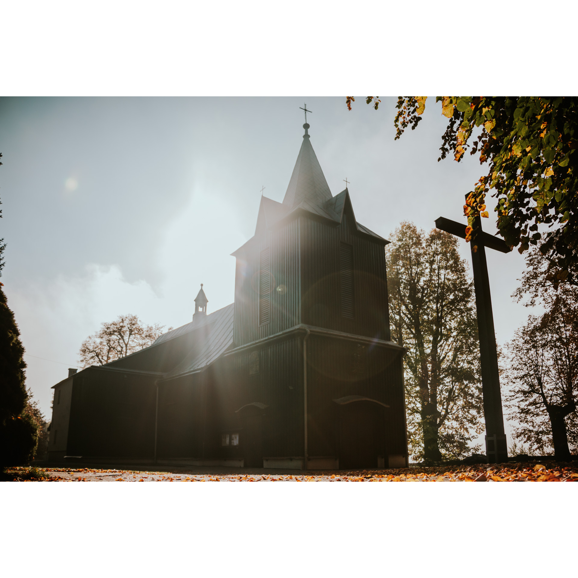 A dark, wooden church with a soaring tower on the front and a tall wooden cross in front of the entrance