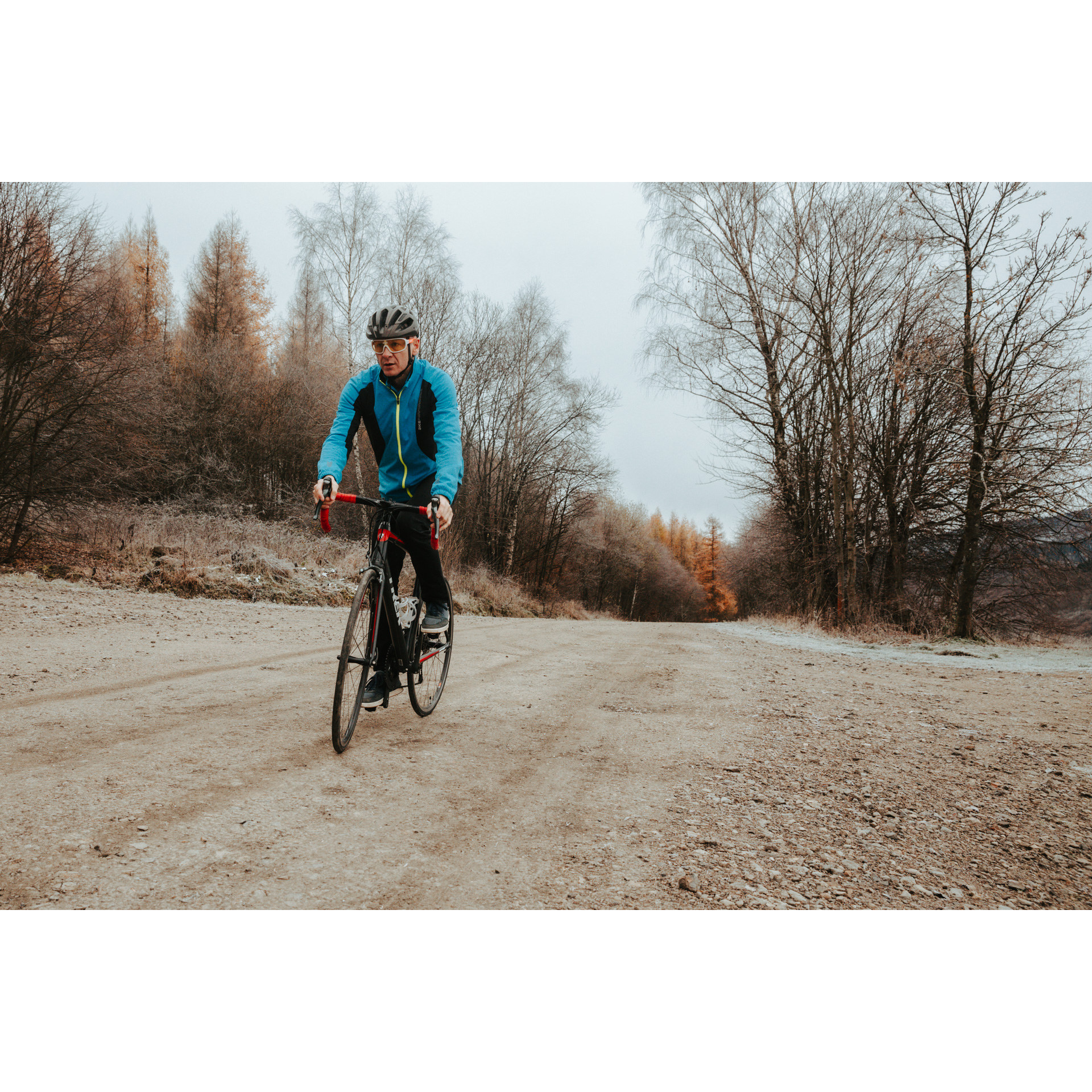 Rowerzysta w niebieskiej kurtce i kasku, jadący na czerwonym rowerze po piaszczystej drodze pod górkę, w tle kolorowe drzewa