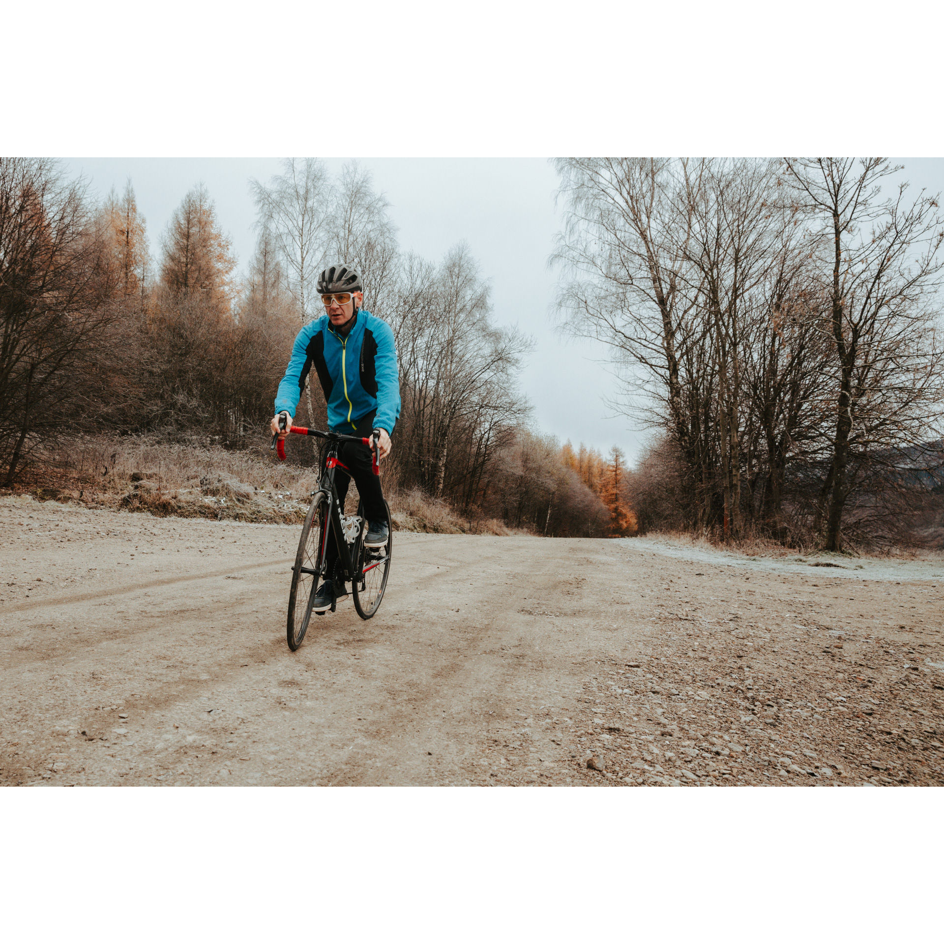 A cyclist in a blue jacket and helmet riding a red bicycle on a sandy road uphill, colorful trees in the background