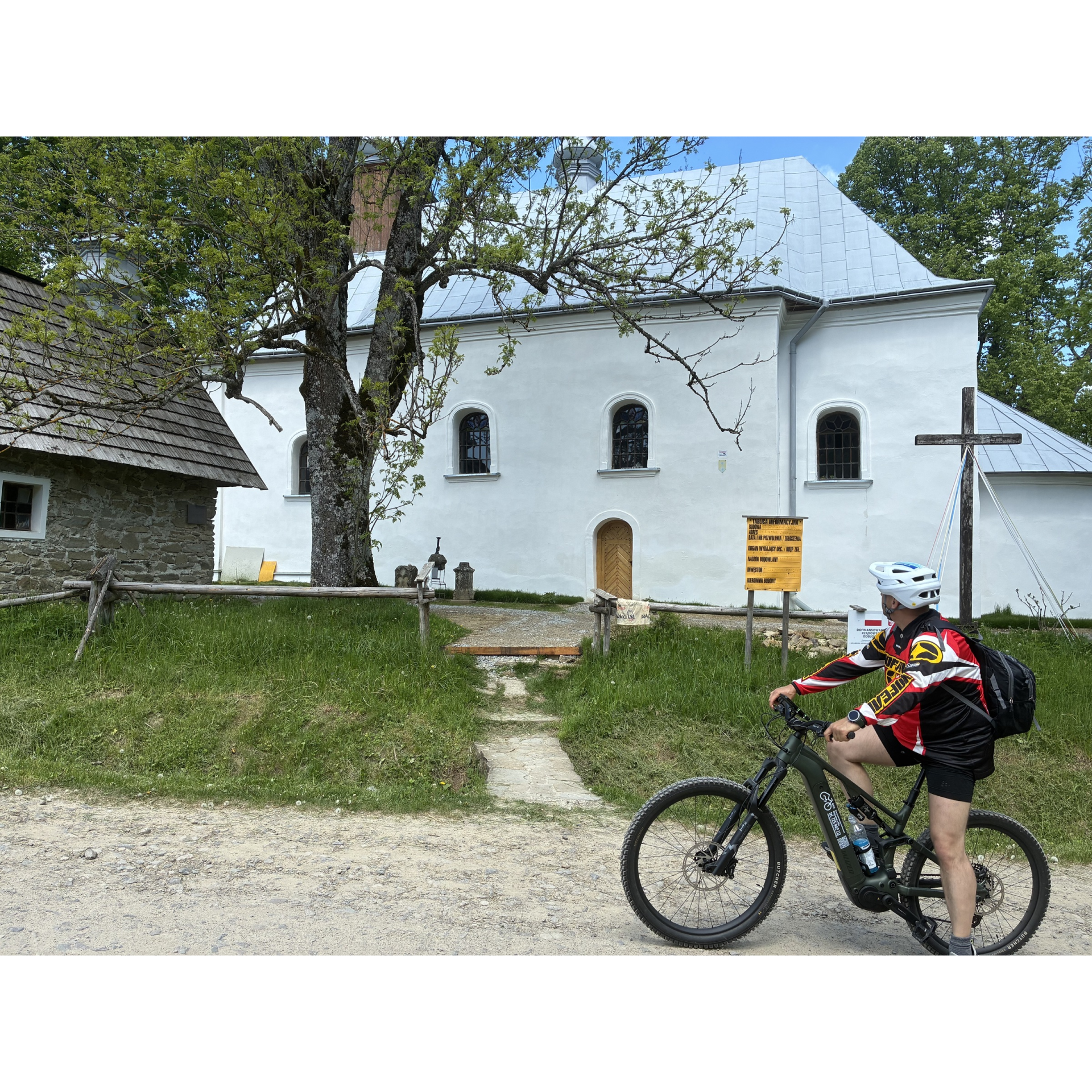 Rowerzysta zatrzymany przy białym wiejskim kościele i drewnianej kapliczce.