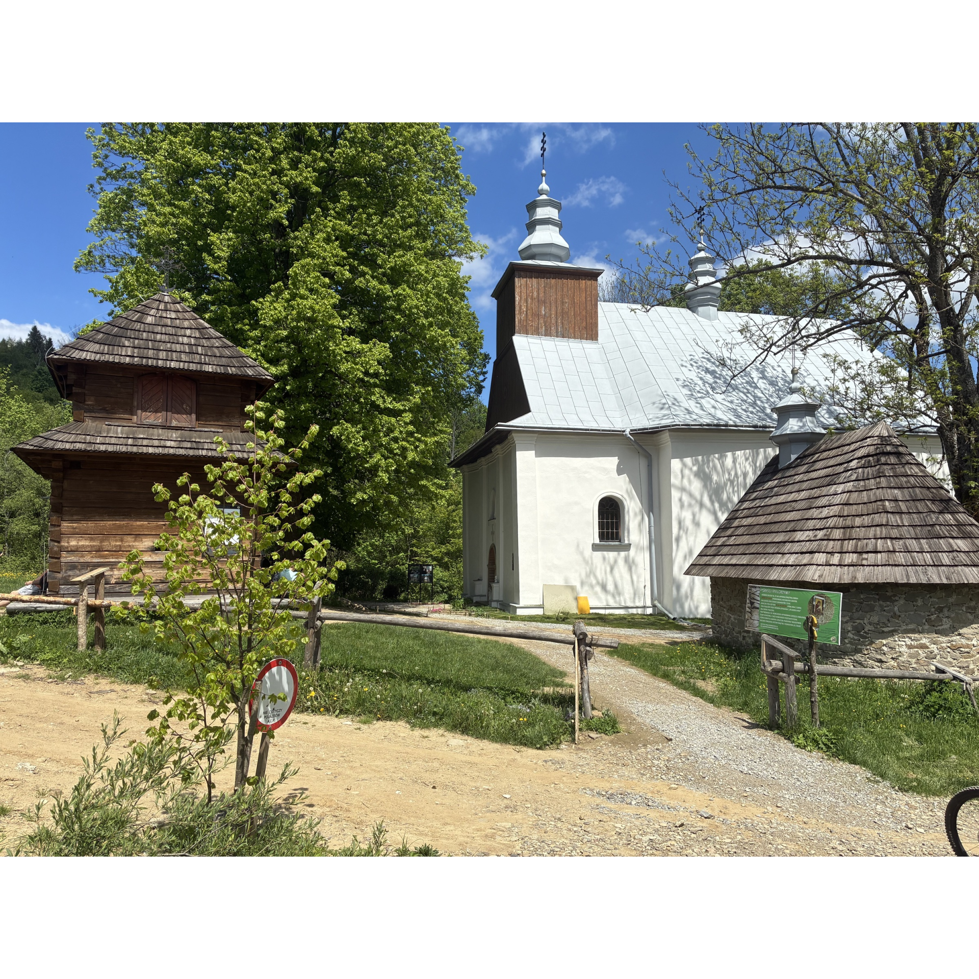 Zespół drewnianych i murowanych budynków sakralnych otoczonych zielenią przy wiejskiej ścieżce.