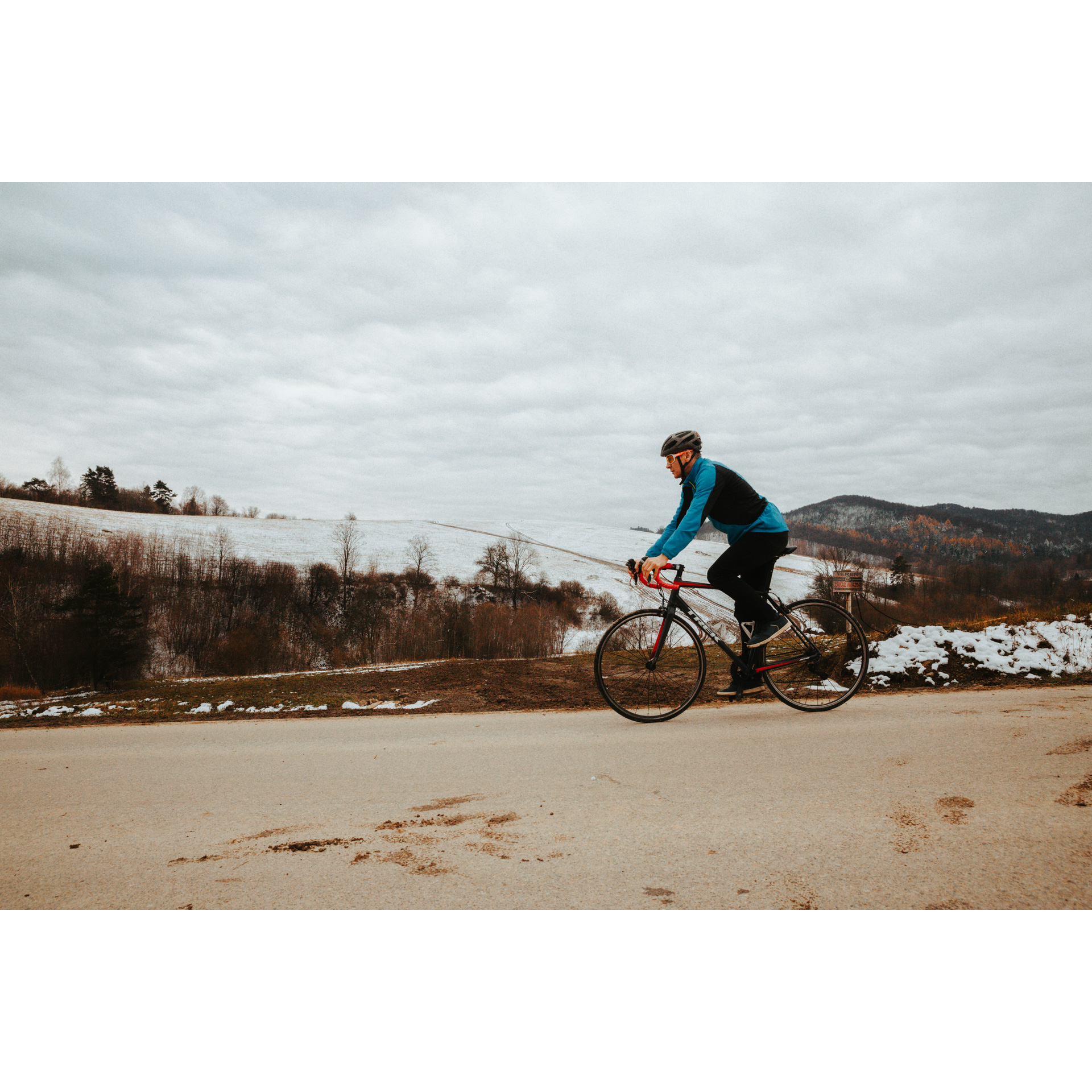 Rowerzysta w kasku i niebieskiej kurtce jadący po asfaltowej drodze na czerwono-czarnym rowerze, w tle zaśnieżone pagórki i brązowo-rude krzewy oraz jasne niebo