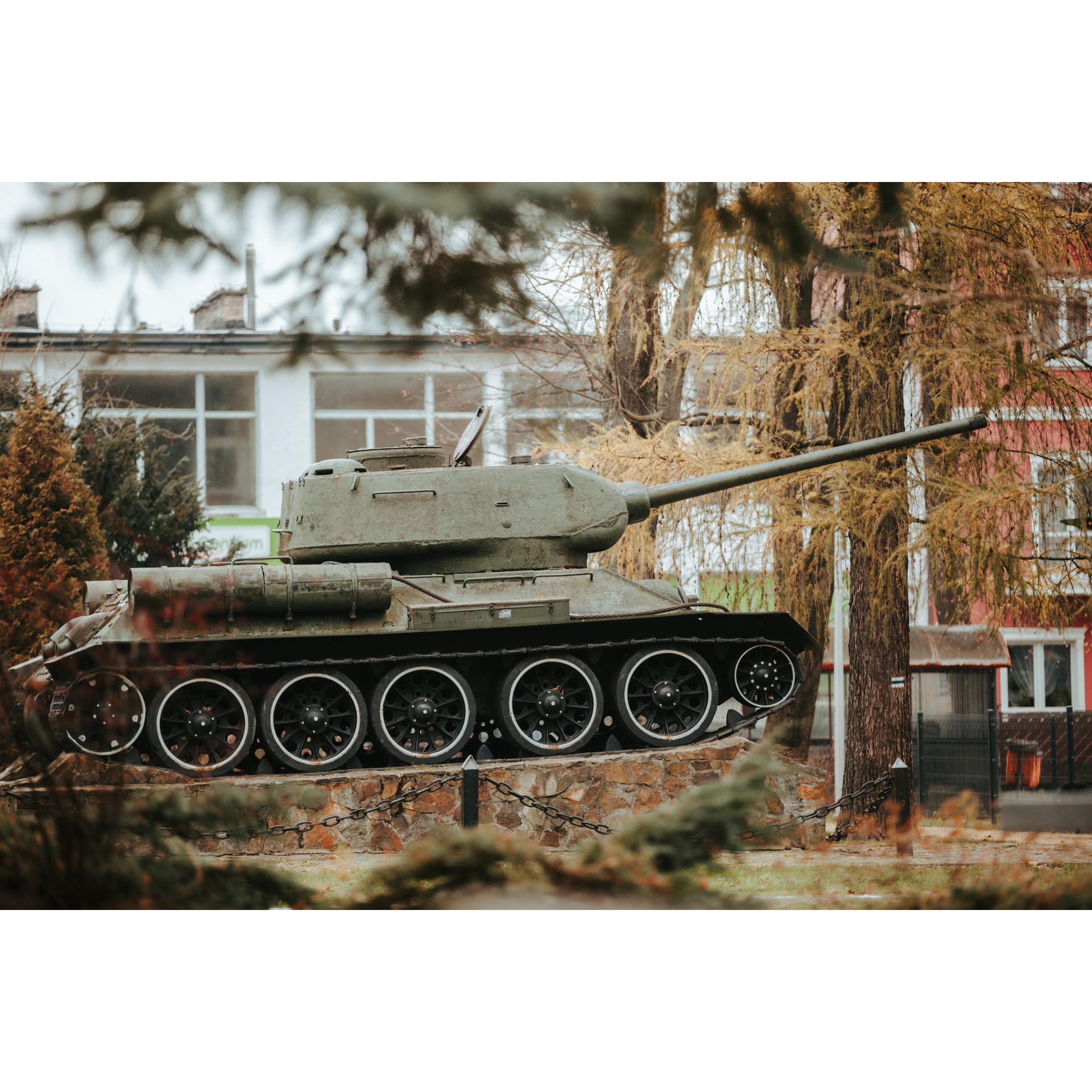 A green, historic tank with the barrel pointing to the right, standing on a brick platform, in the background a building with large windows