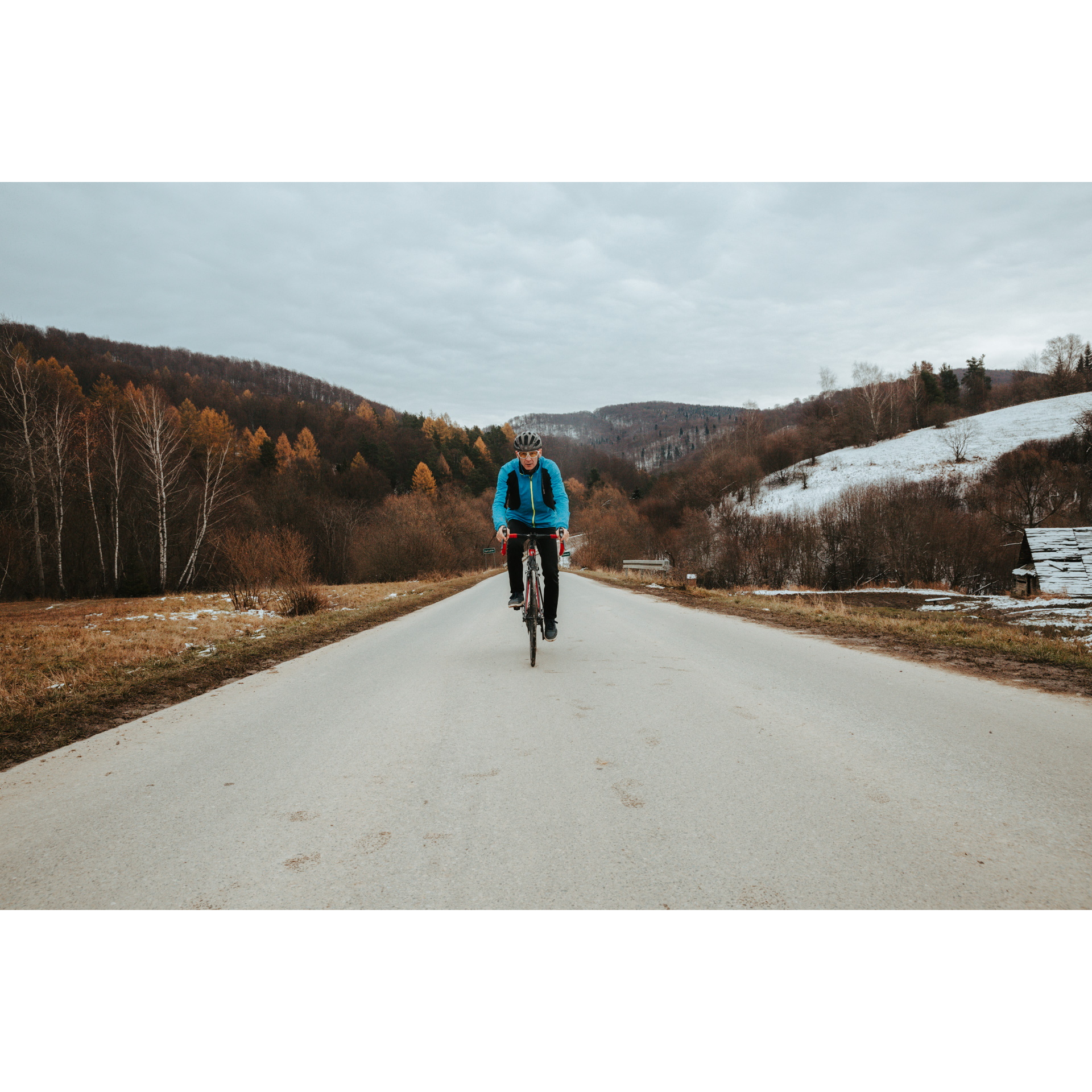 A cyclist in a helmet, glasses and a blue jacket riding straight ahead on an asphalt road on a red bicycle, snowy hills on the right, hills covered with brown-red trees on the left
