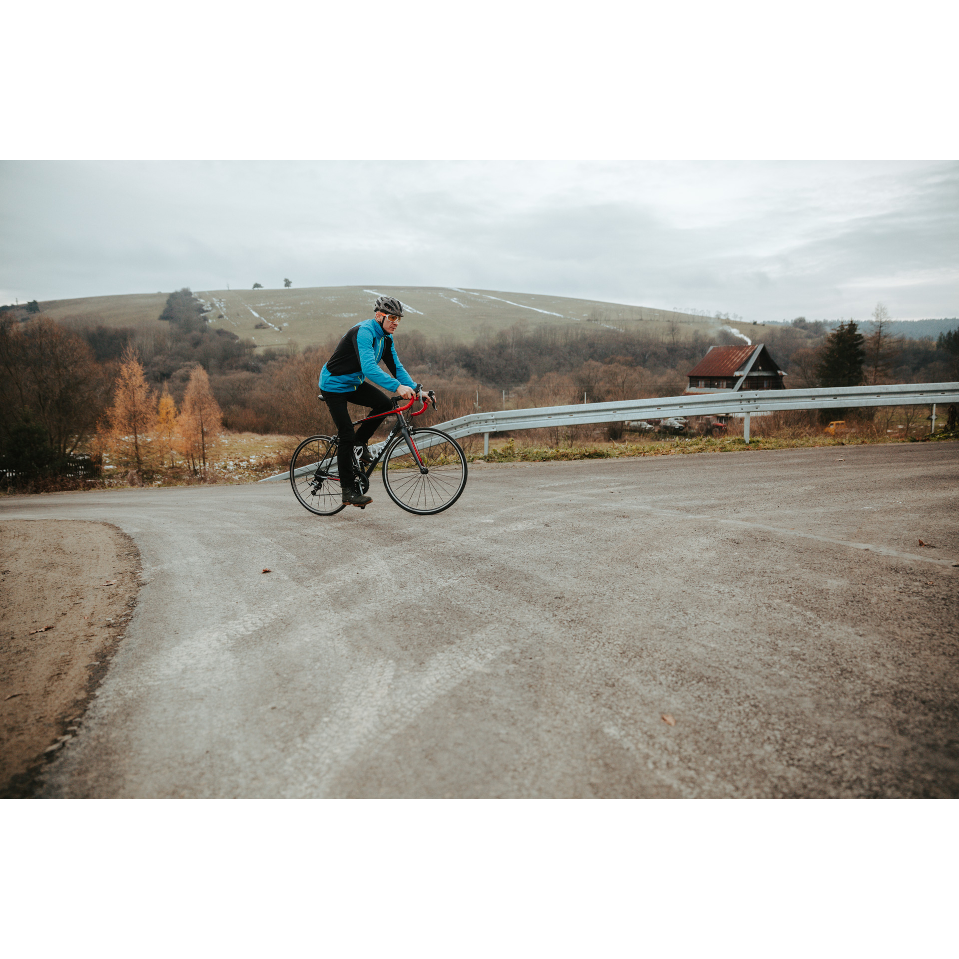 Rowerzysta w niebieskiej kurtce i kasku na czerwonym rowerze, jadący pod górkę na asfaltowej drodze i skręcający w prawo, w tle wzgórza i drzewa