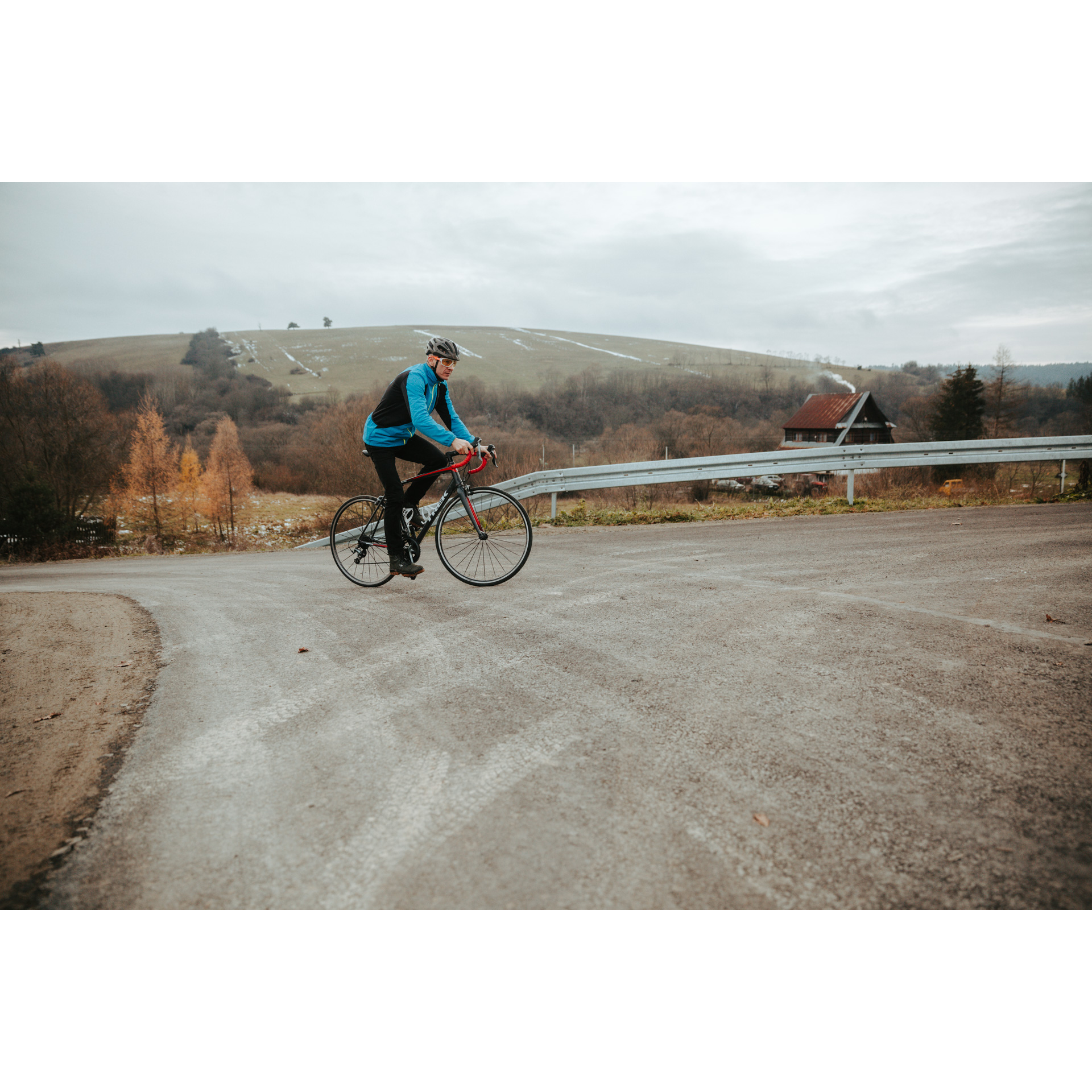 A cyclist in a blue jacket and helmet on a red bicycle going uphill on an asphalt road and turning right, hills and trees in the background