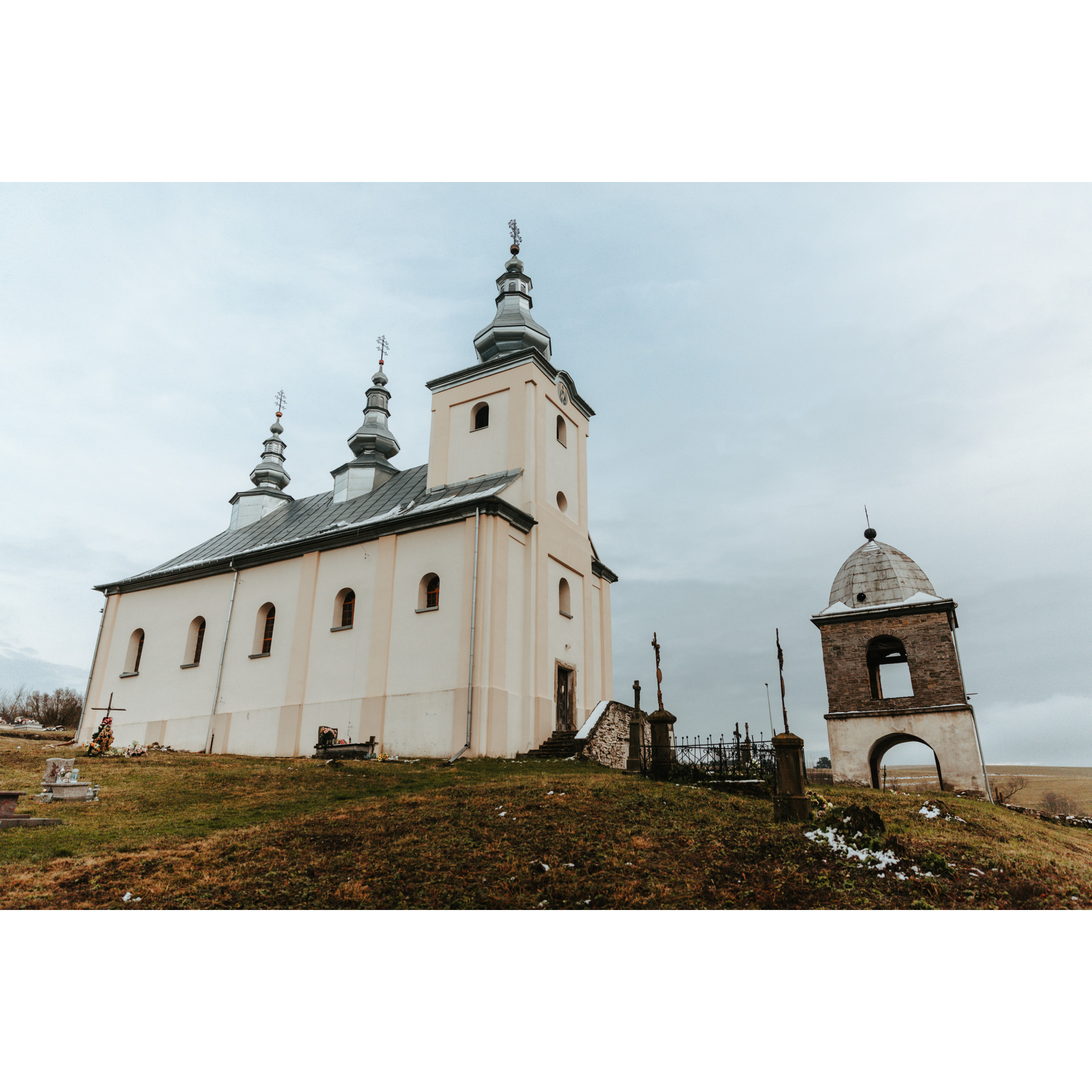 Biała, murowana cerkiew z szarym dachem zwieńczonym trzema kopułami i krzyżami, obok niska dzwonnica z kopulastym dachem i krzyżem
