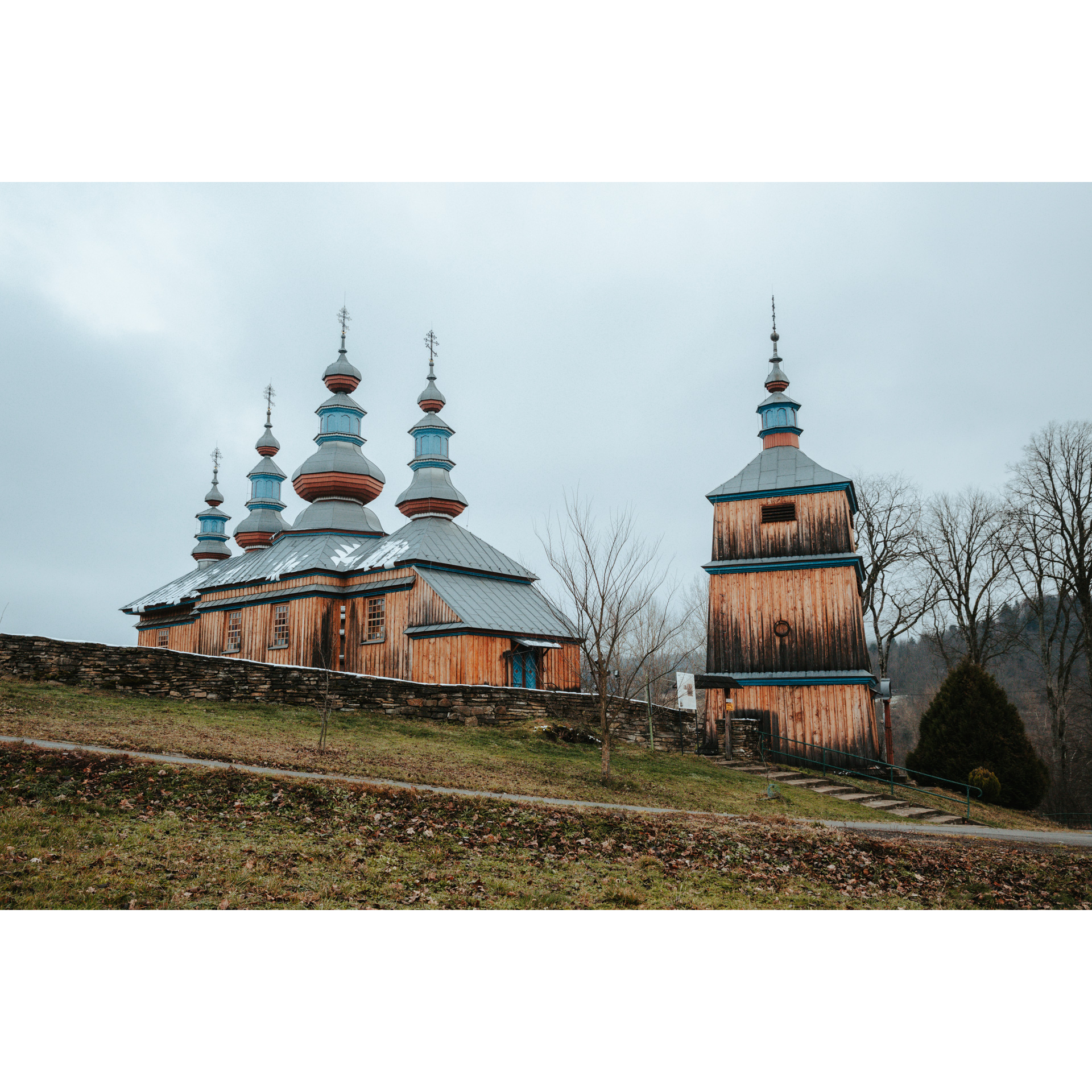 A wooden, historic Orthodox church topped with a roof with four domes and crosses, next to it there is a wooden belfry topped with a roof with one dome and a cross