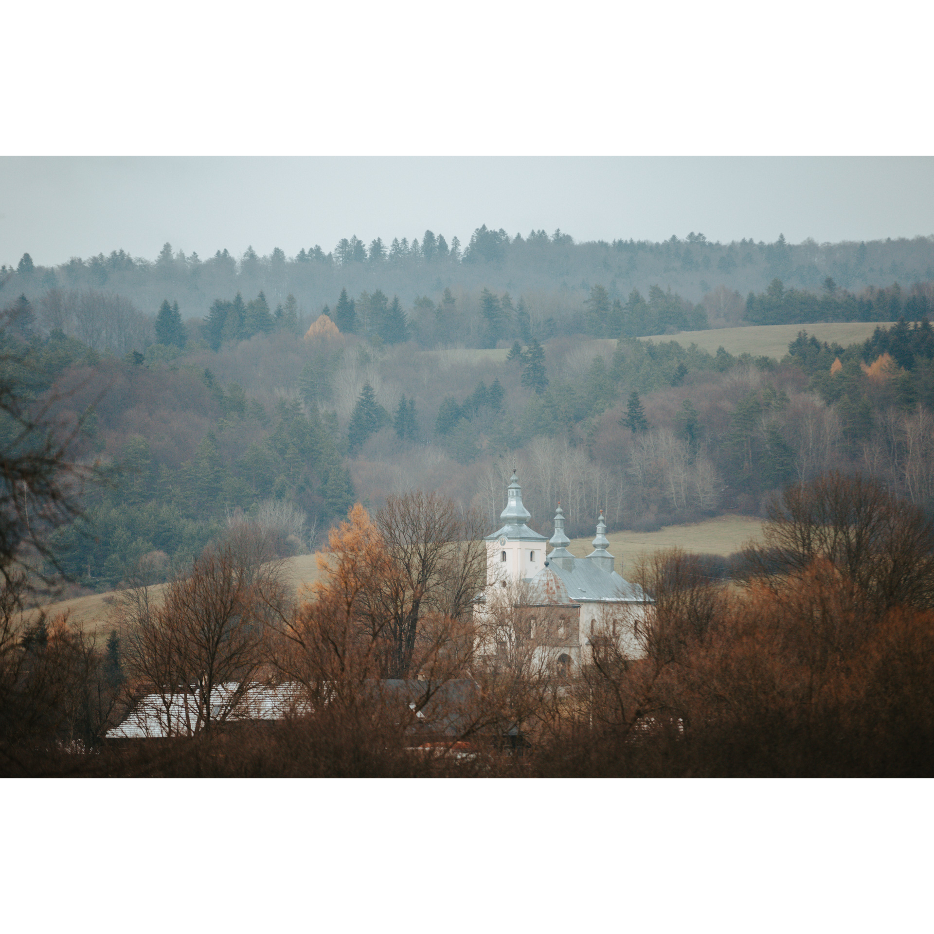 A white, brick Orthodox church in the distance among brown trees, in the background hills covered with colorful trees