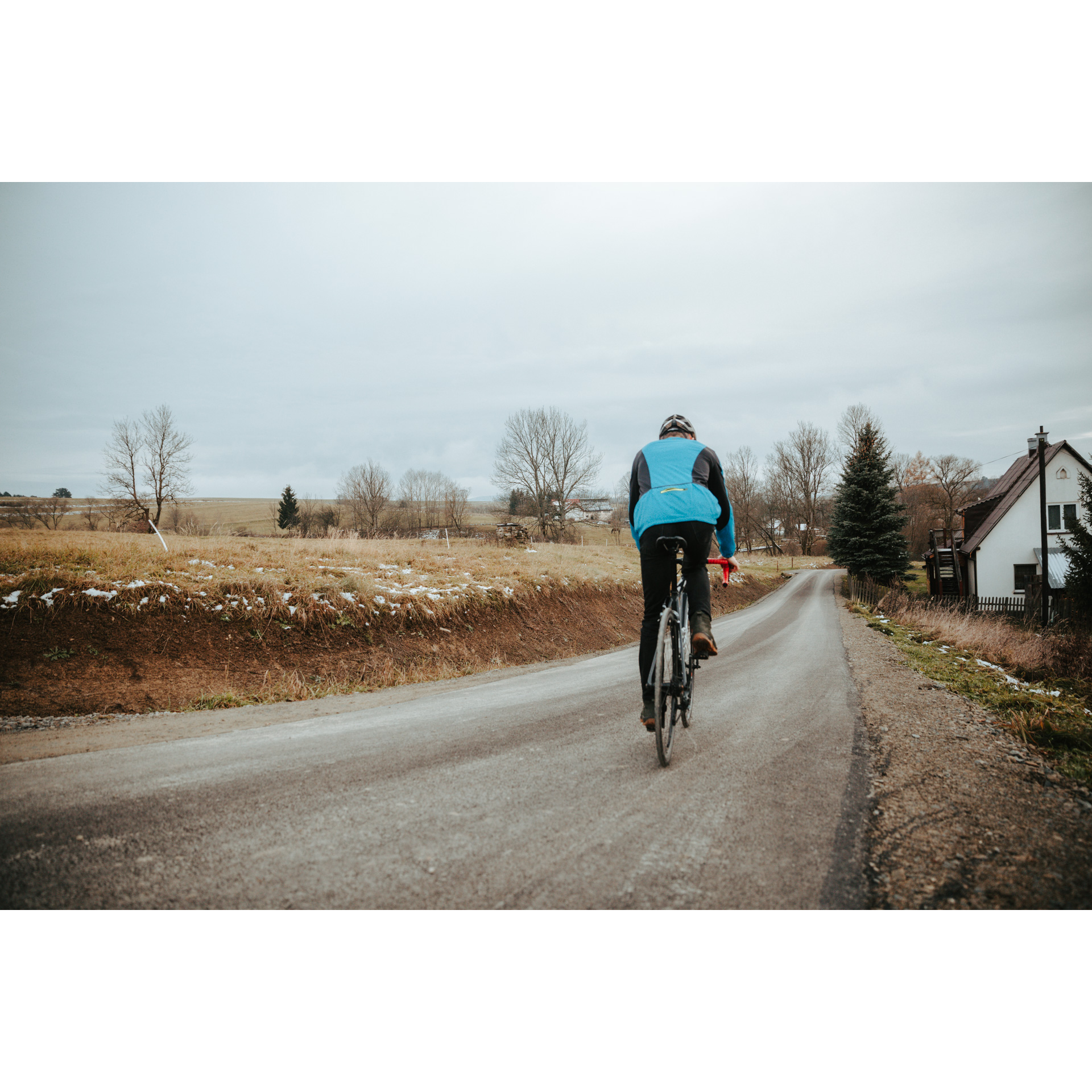 A cyclist in a blue jacket seen from the back, cycling down a hill on an asphalt road, on the right a white house with a triangular roof, on the left slightly snowy fields