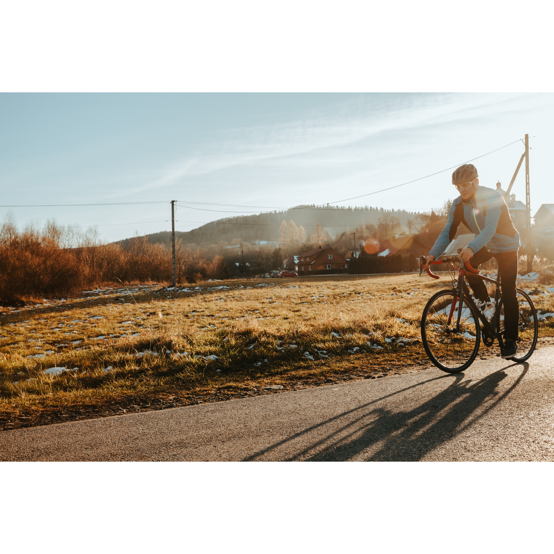 Rowerzysta w niebieskiej kurtce i kasku jadący asfaltową drogą na czerwono-czarnym rowerze, w tle lekko zaśnieżona polana, krzewy, budynki gospodarcze i wzgórze