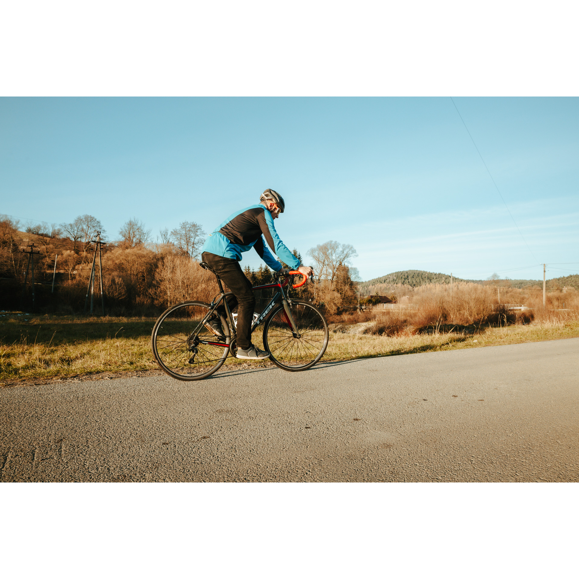 Rowerzysta w niebieskiej kurtce na czerwono-czarnym rowerze, jadący w prawą stronę po asfaltowej drodze, w tle brązowe krzewy i błękitne niebo