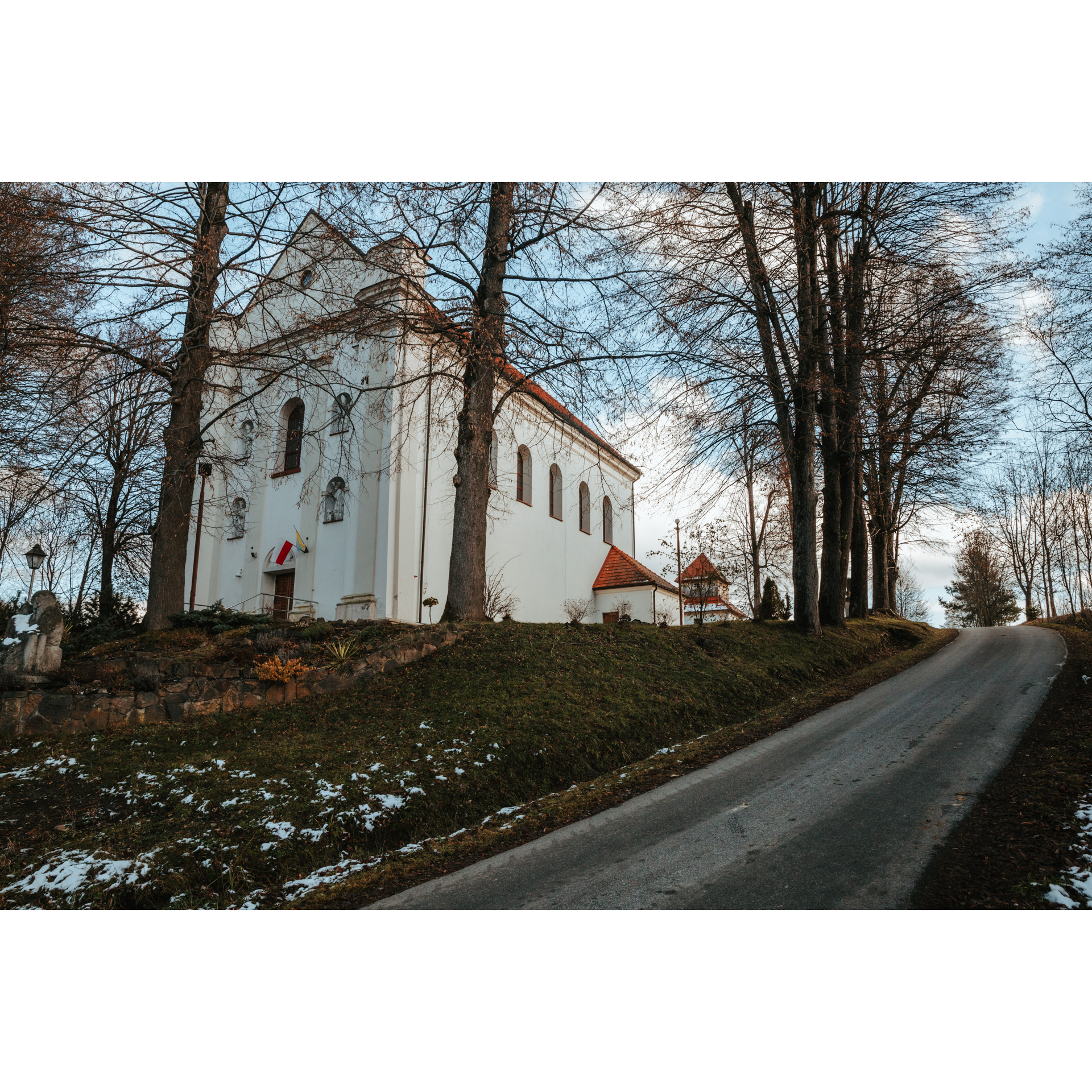 Standing behind leafless trees, a white, brick, rectangular church on top of a hill, accessed by an asphalt path