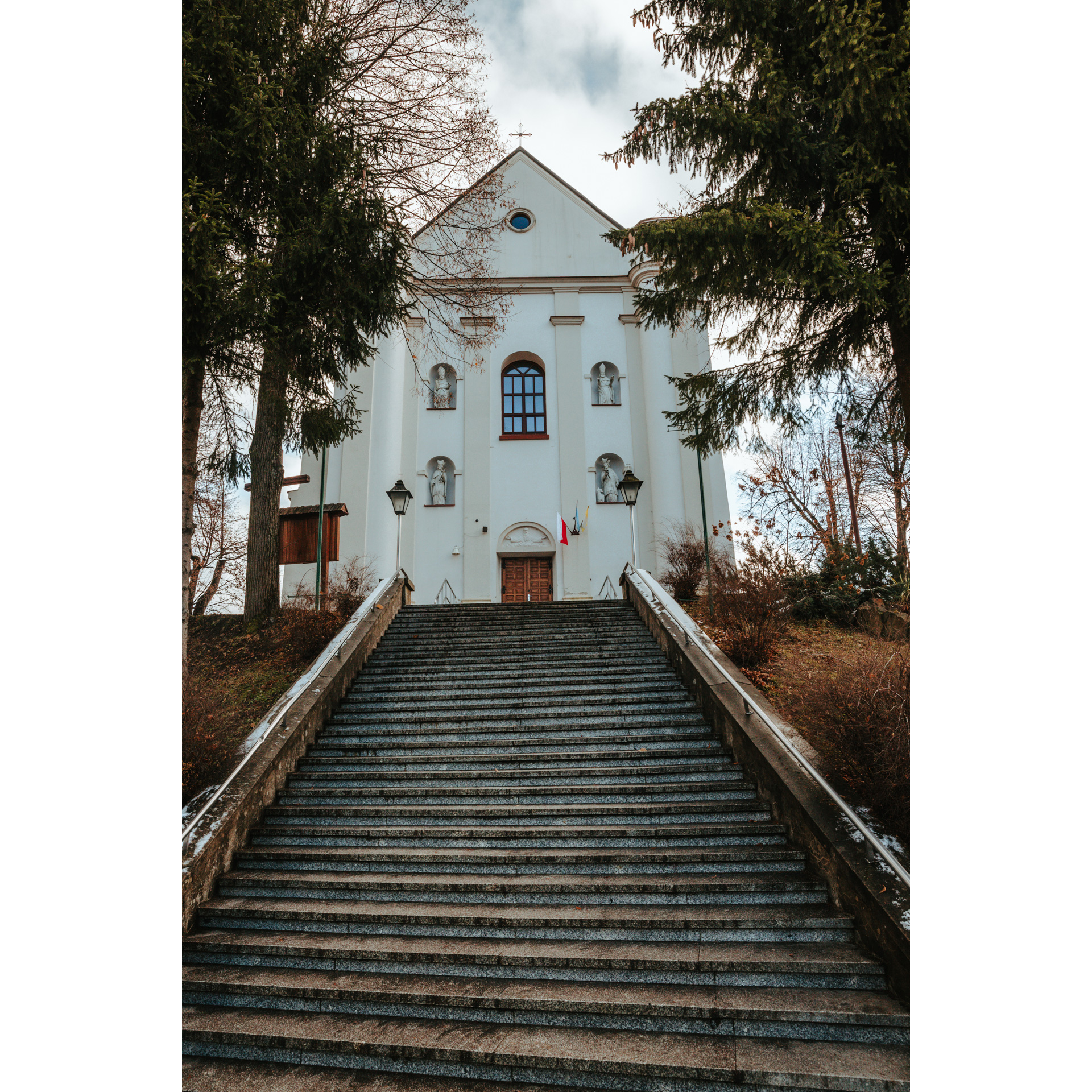 Biały, murowany kościół na szczycie schodów z czterema wnękami z figurami świętych w fasadzie, półokrągłym oknem, drzwiami i krzyżem na szczycie