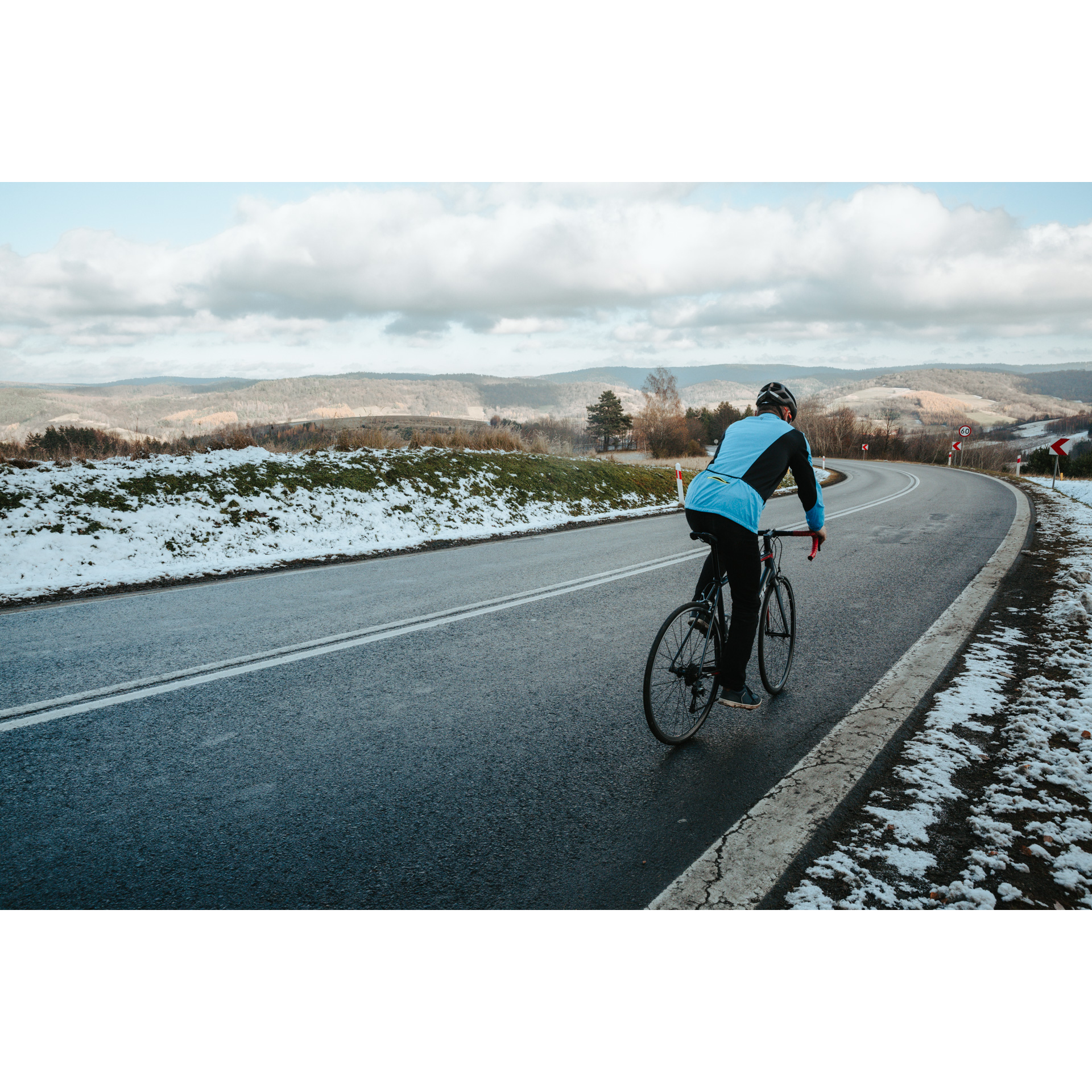 A cyclist in a blue jacket cycling down a two-lane asphalt road turning left, snowy fields in the background, mountains and gray sky in the distance
