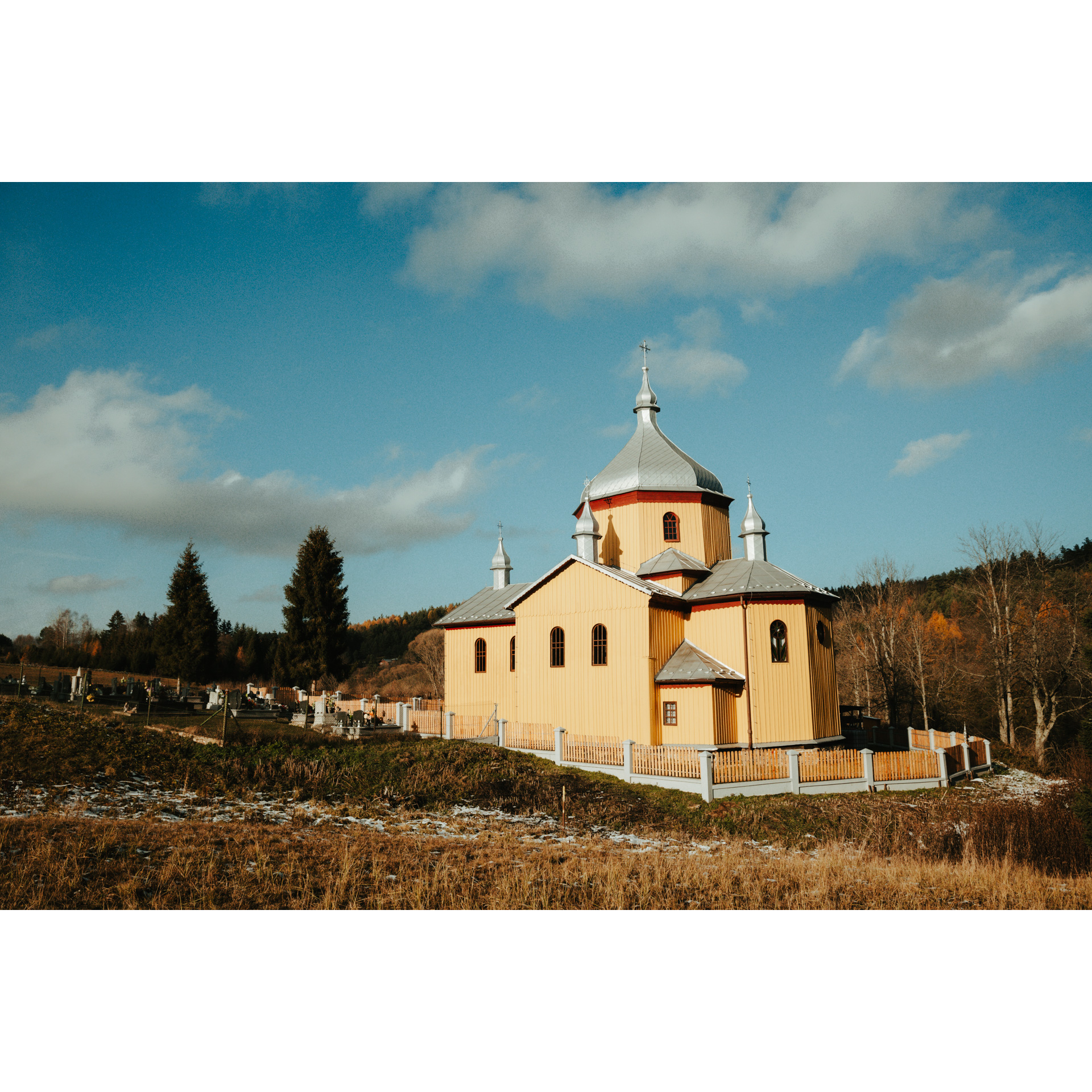 A wooden church surrounded by a fence, topped with a silver roof with 2 domes and crosses, a cemetery next to it, trees, hills, blue sky and clouds in the background