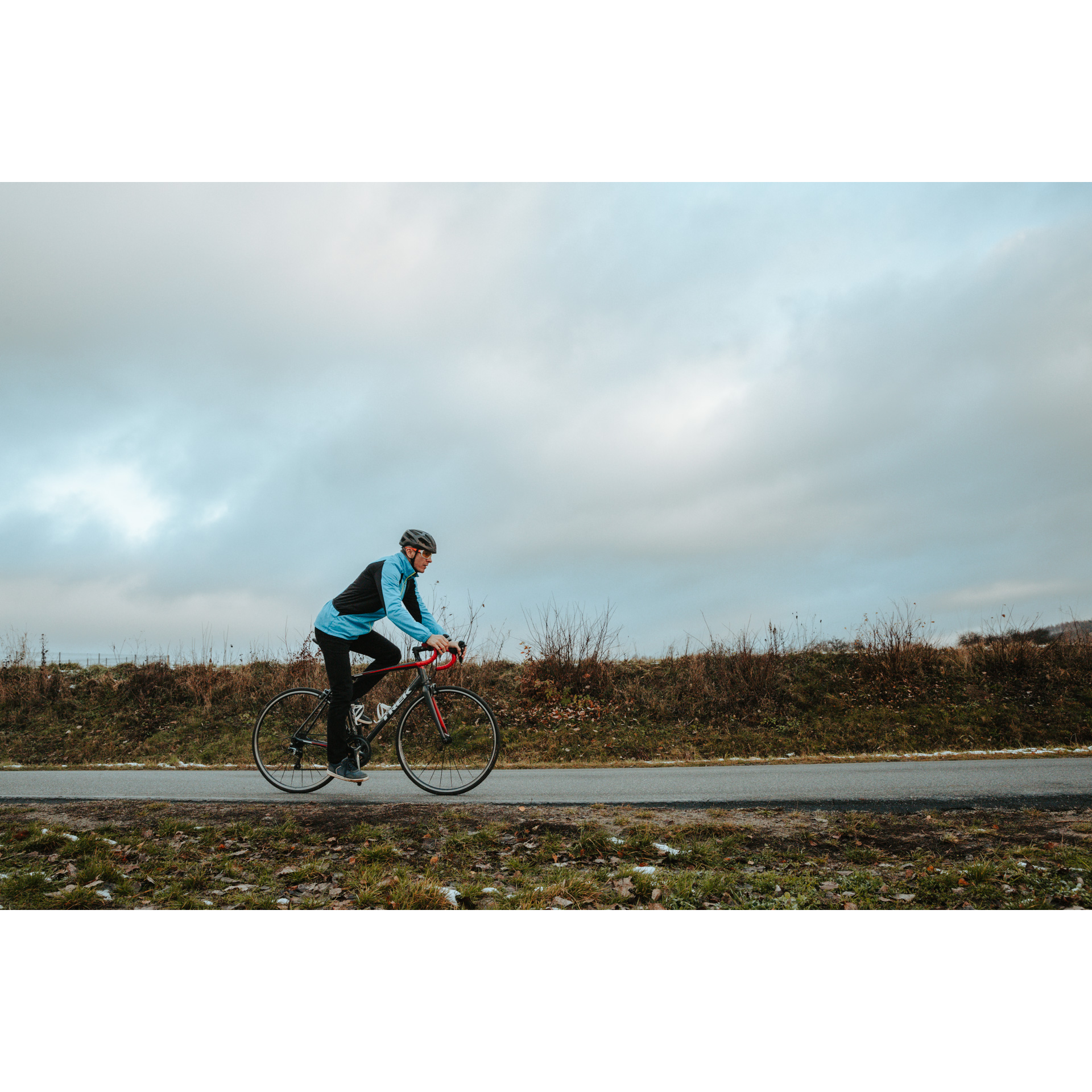 Rowerzysta w niebieskiej kurtce, czarnych spodniach i kasku, jadący na czerwono-czarnym rowerze po asfaltowej drodze, w tle zachmurzone niebo