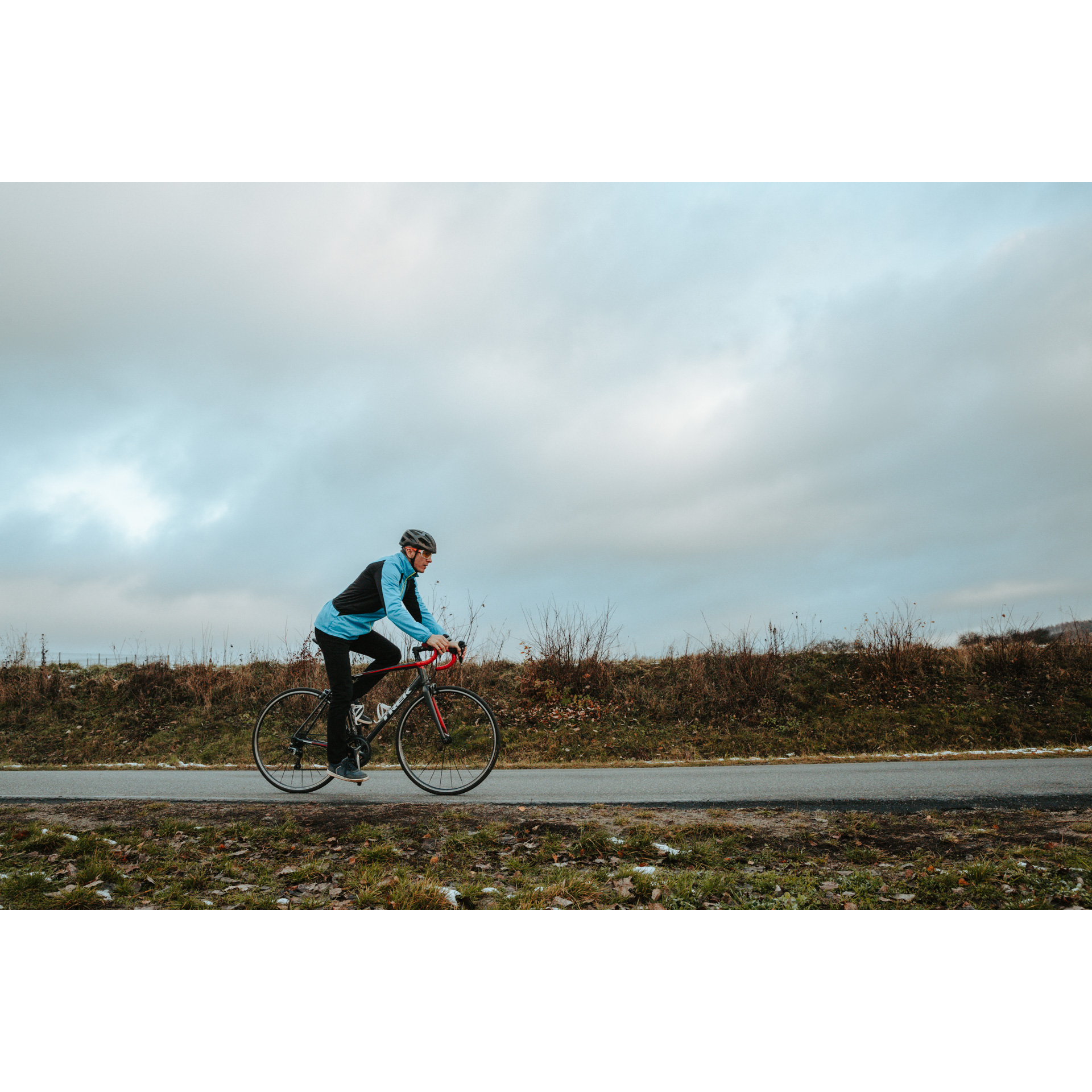 A cyclist in a blue jacket, black pants and a helmet, riding a red and black bicycle on an asphalt road, cloudy sky in the background