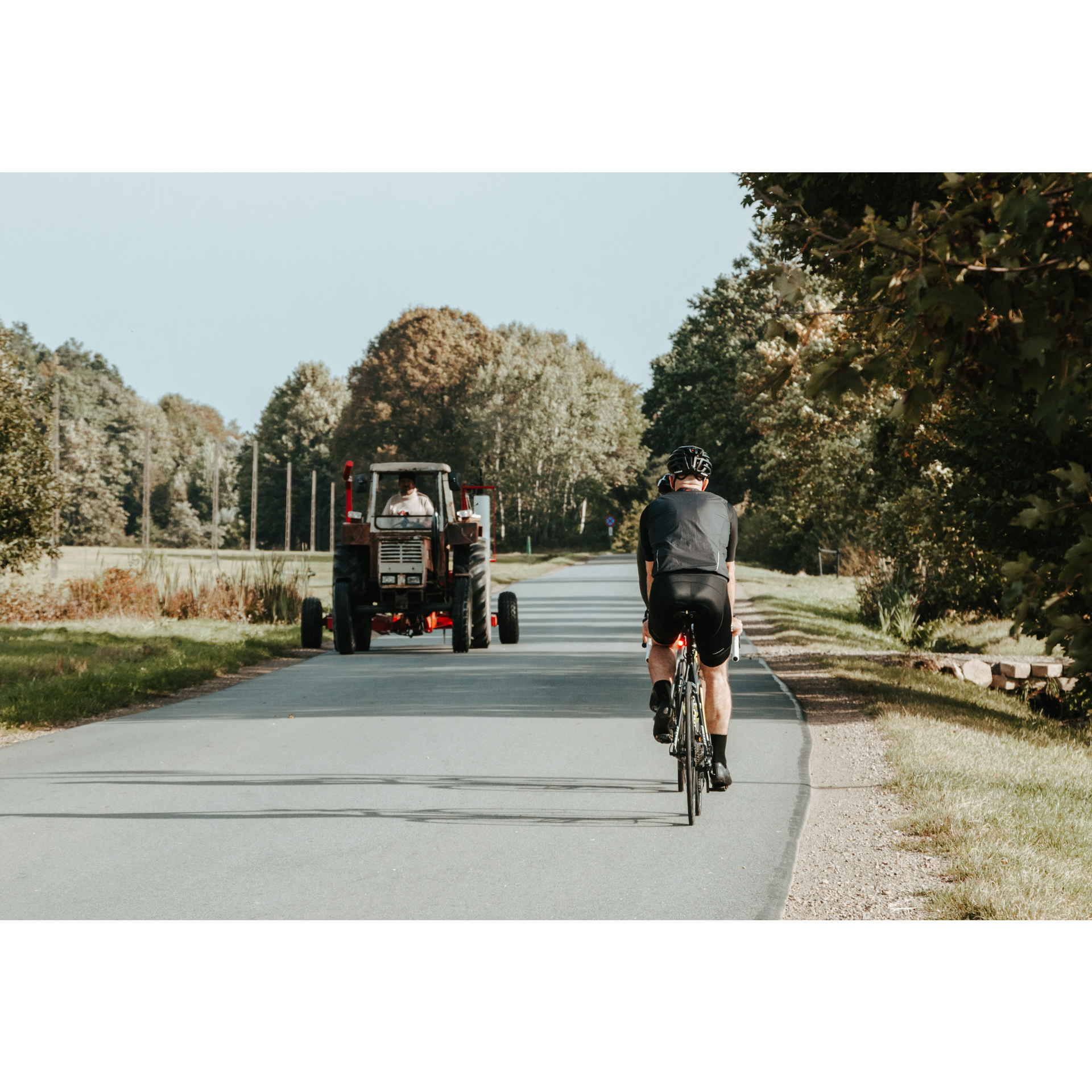 Dwóch kolarzy na rowerach w kaskach i czarnych strojach jadących asfaltową drogą wymijających się z traktorem jadącym w przeciwnym kierunku, w tle drzewa