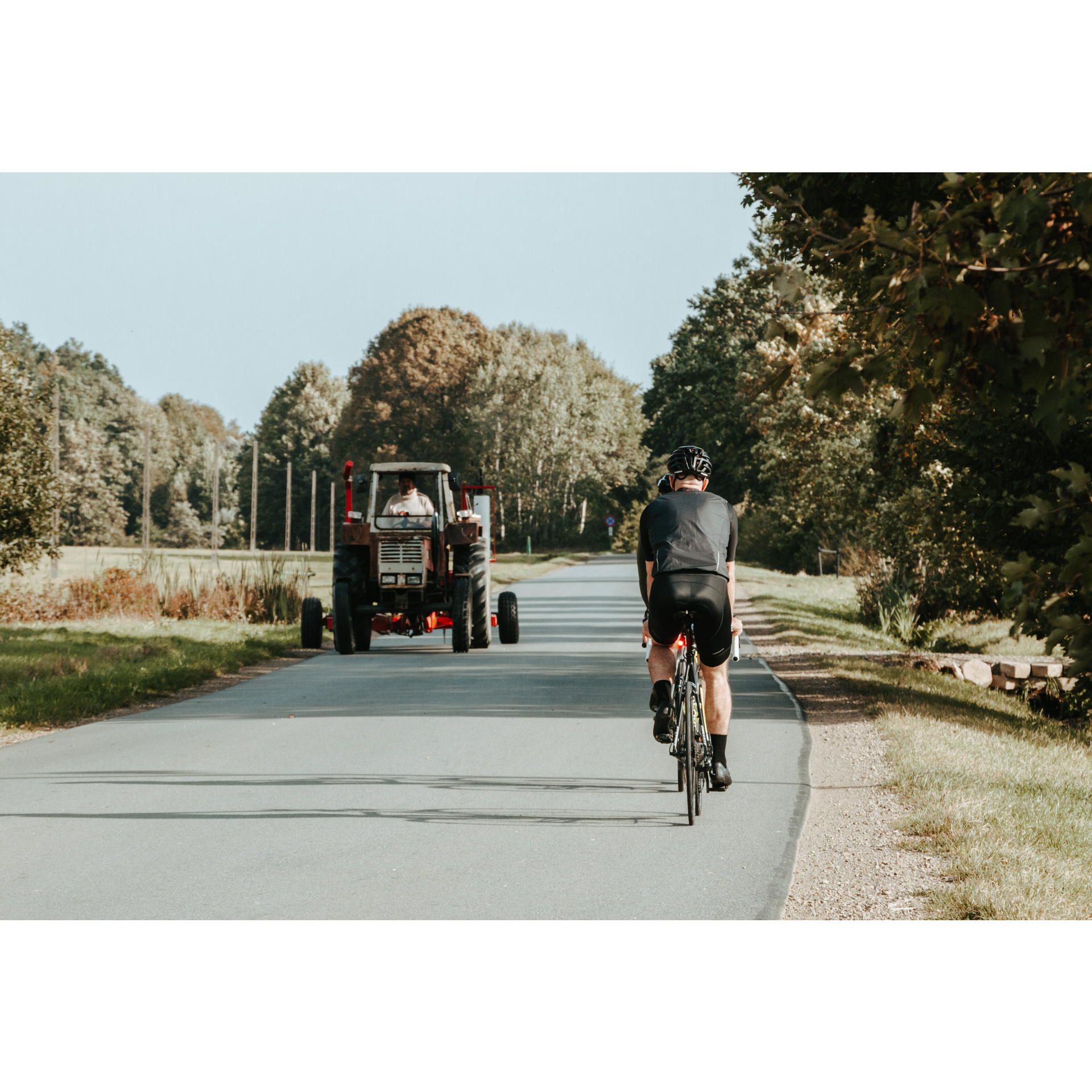 Two cyclists on bicycles in helmets and black clothes riding on an asphalt road passing each other with a tractor going in the opposite direction, trees in the background