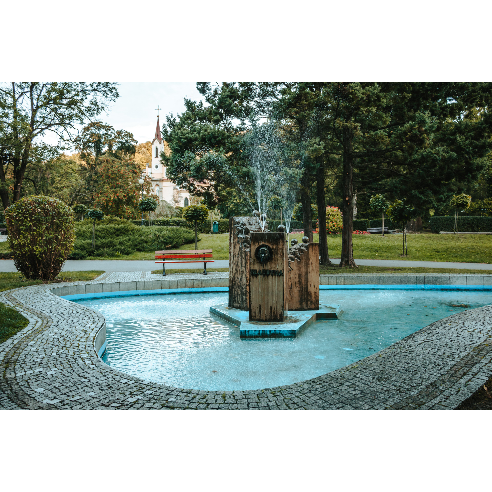 An irregularly shaped fountain surrounded by a cube wall and gushing water from three rectangular elements in the center, trees and a soaring white church in the background