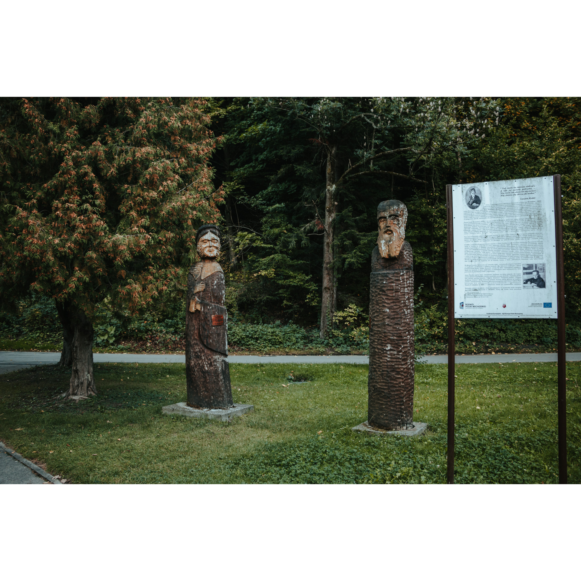 Tablica informacyjna i dwie figury drewniane przedstawiające mężczyznę i kobietę, stojące na zielonej trawie wśród drzew 