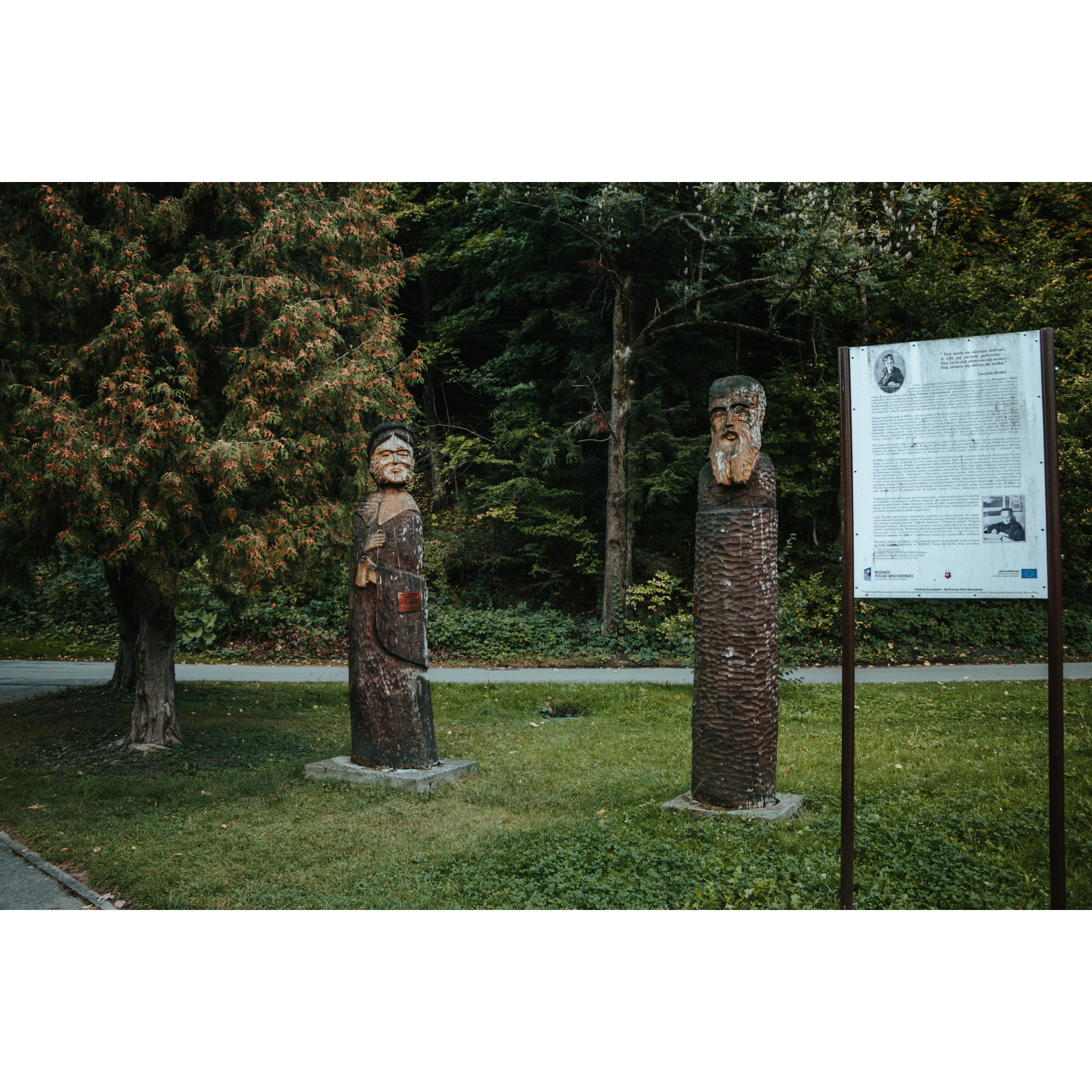 An information board and two wooden figures of a man and a woman standing on the green grass among the trees