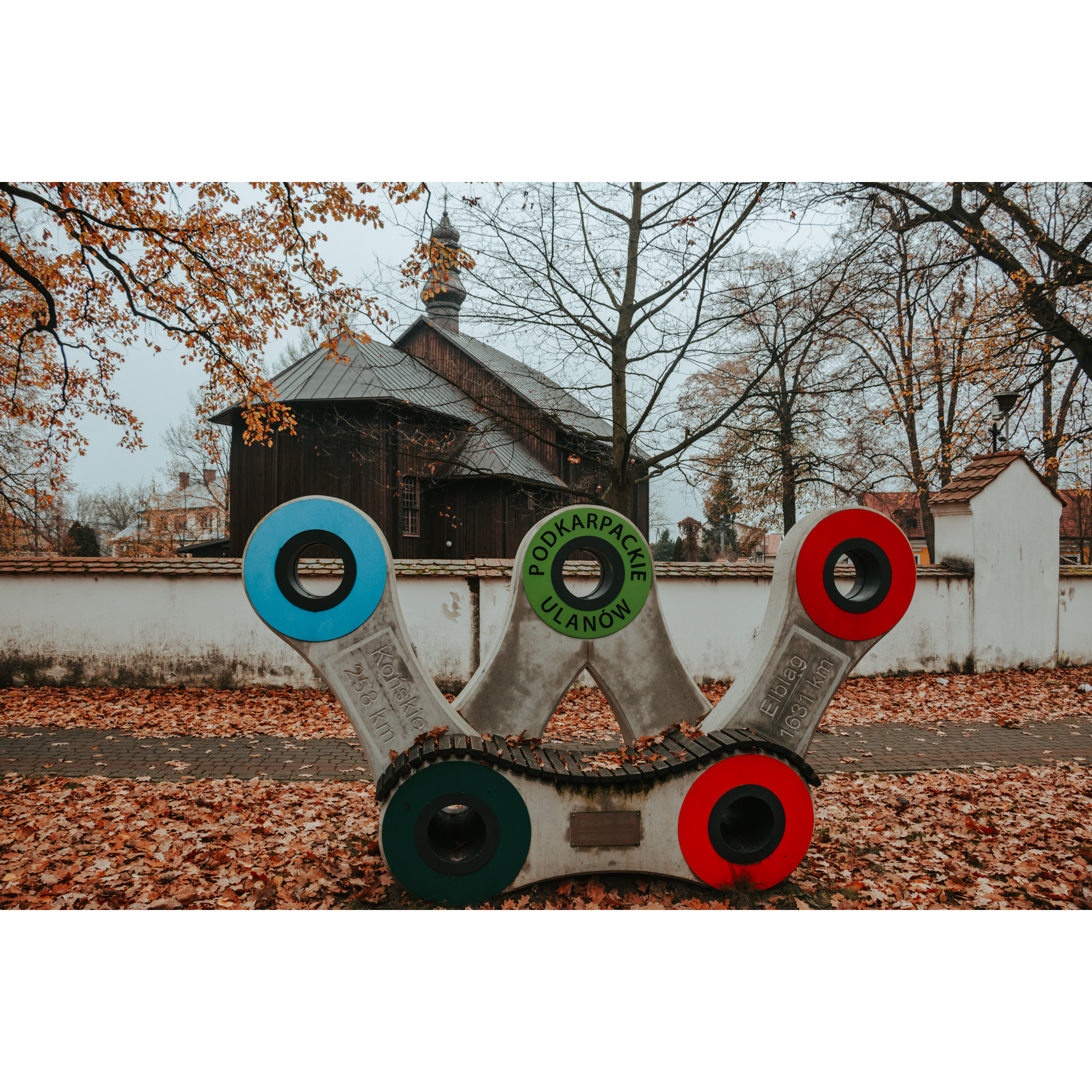 A memorial bench in the shape of a bicycle chain with colorful links, a white wall and a wooden church in the background