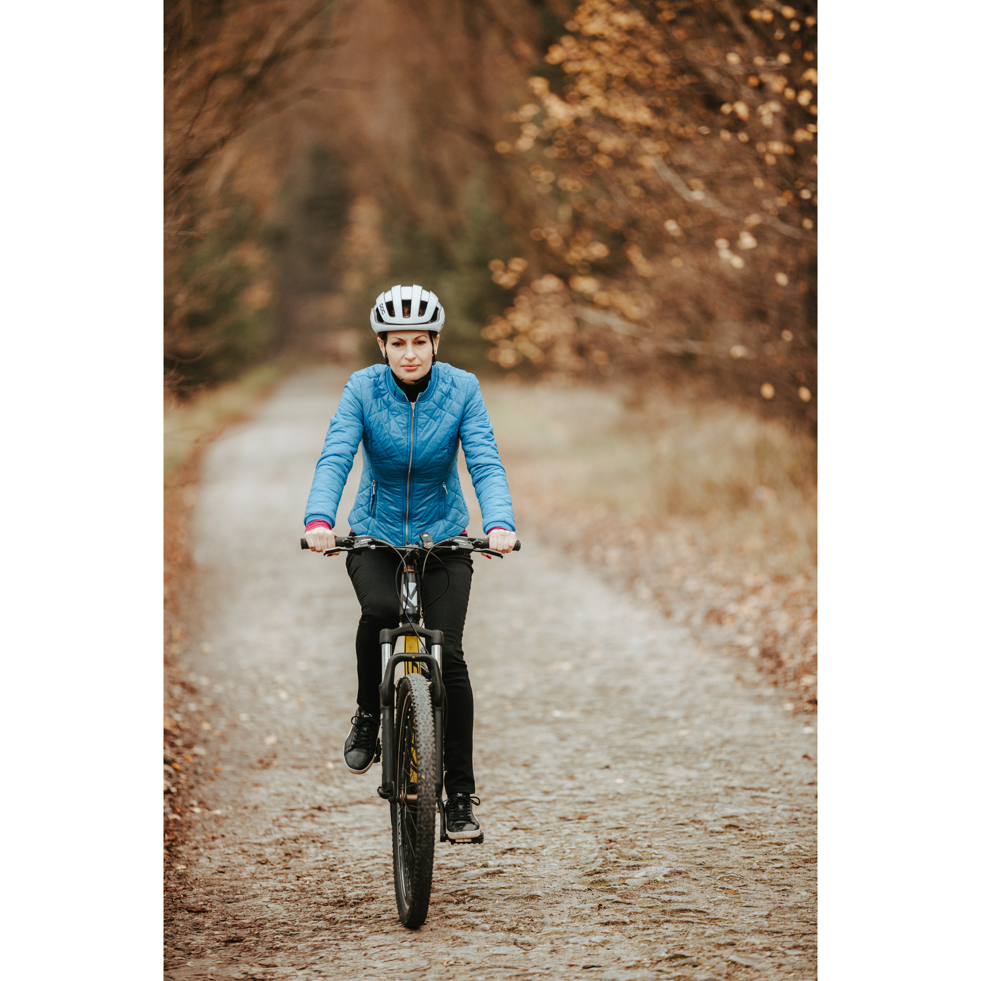 Rowerzystka w niebieskiej kurtce, czarnych spodniach i białym kasku jadąca na rowerze piaszczystą drogą leśną