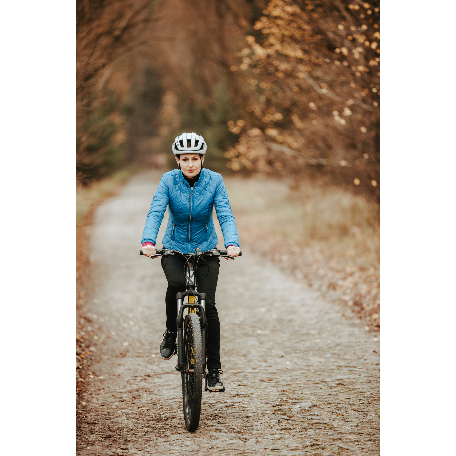 A cyclist in a blue jacket, black pants and a white helmet cycling along a sandy forest road