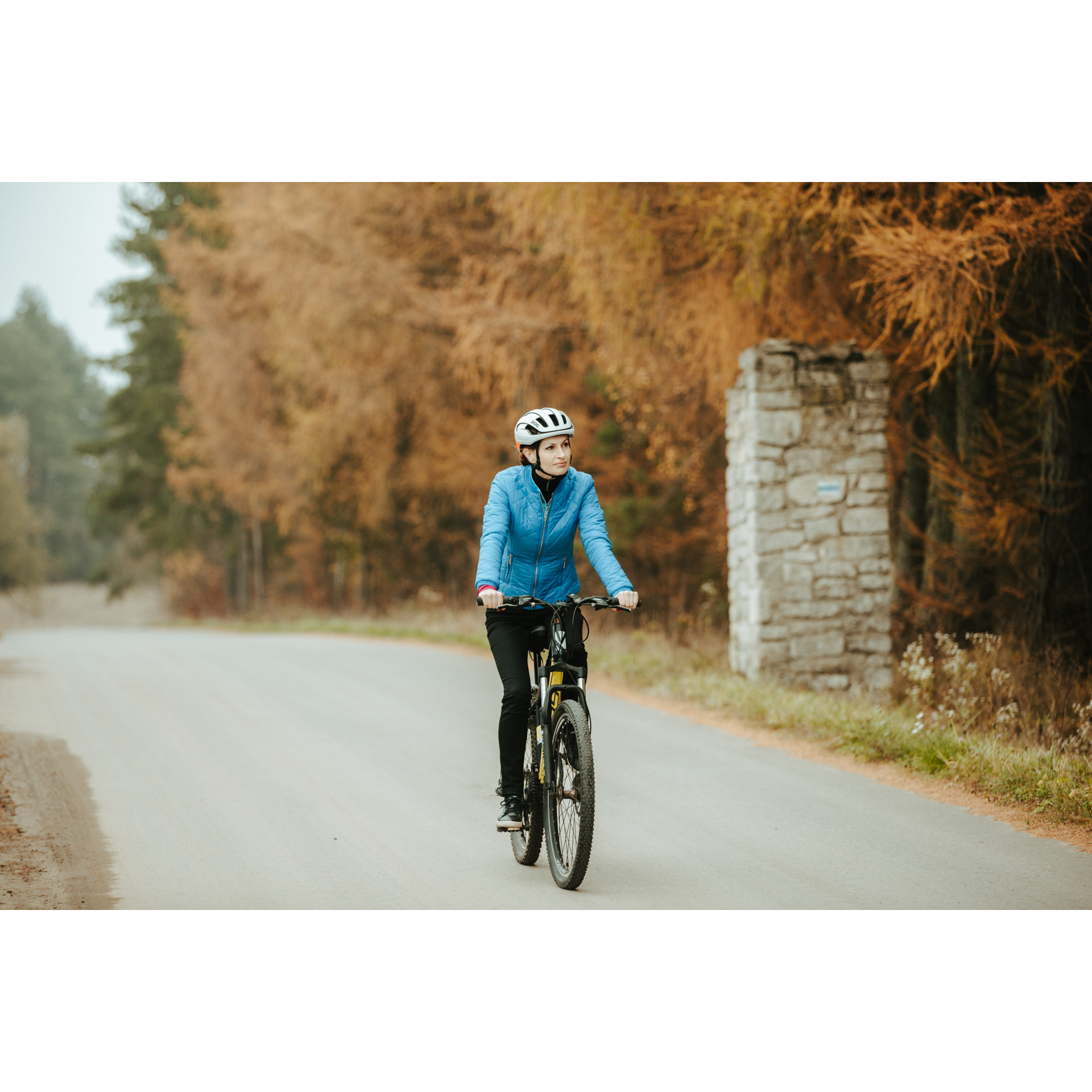 Rowerzystka w niebieskiej kurtce i białym kasku jadąca asfaltową drogą, mijająca brązowe drzewa i murowany słup