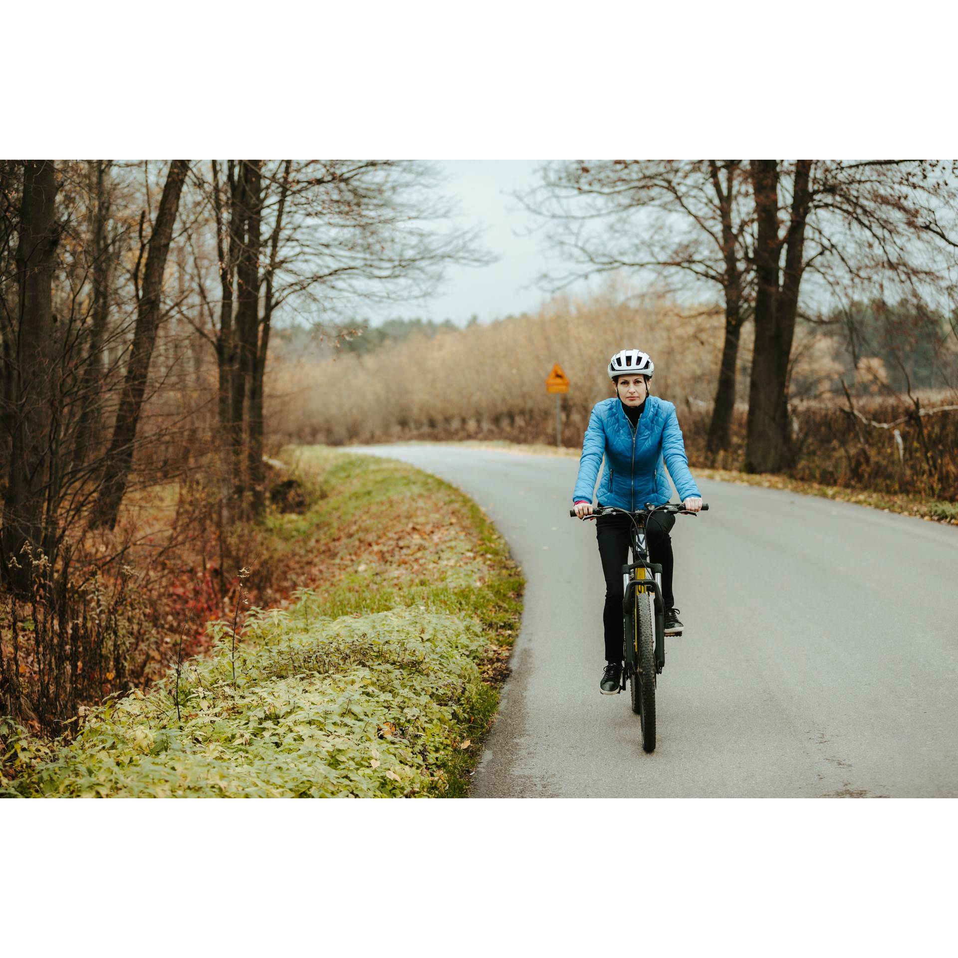 Rowerzystka w niebieskiej kurtce i białym kasku jadąca na rowerze asfaltową drogą, mijająca kolorowe drzewa i krzewy