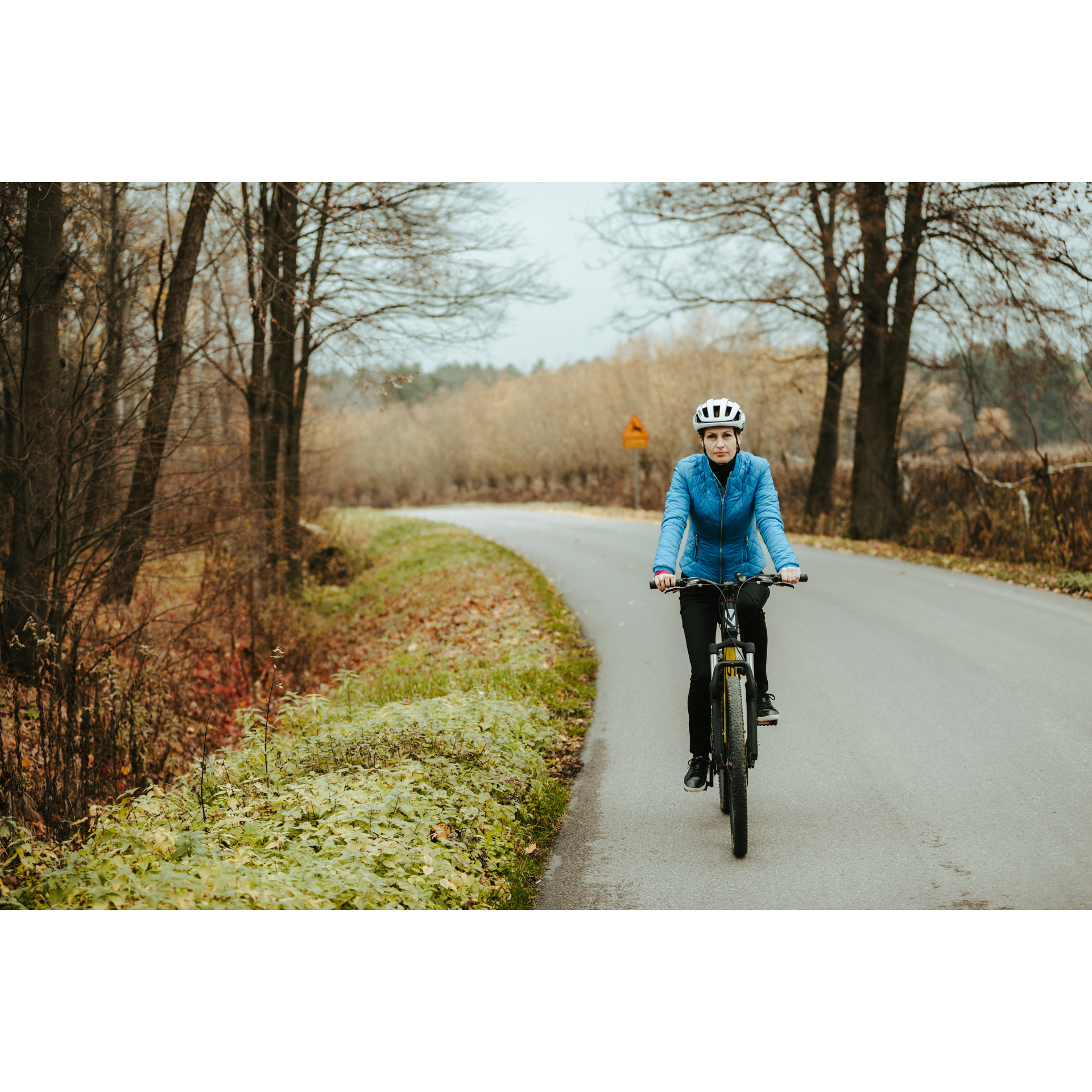 A cyclist in a blue jacket and a white helmet cycling along an asphalt road, passing colorful trees and bushes