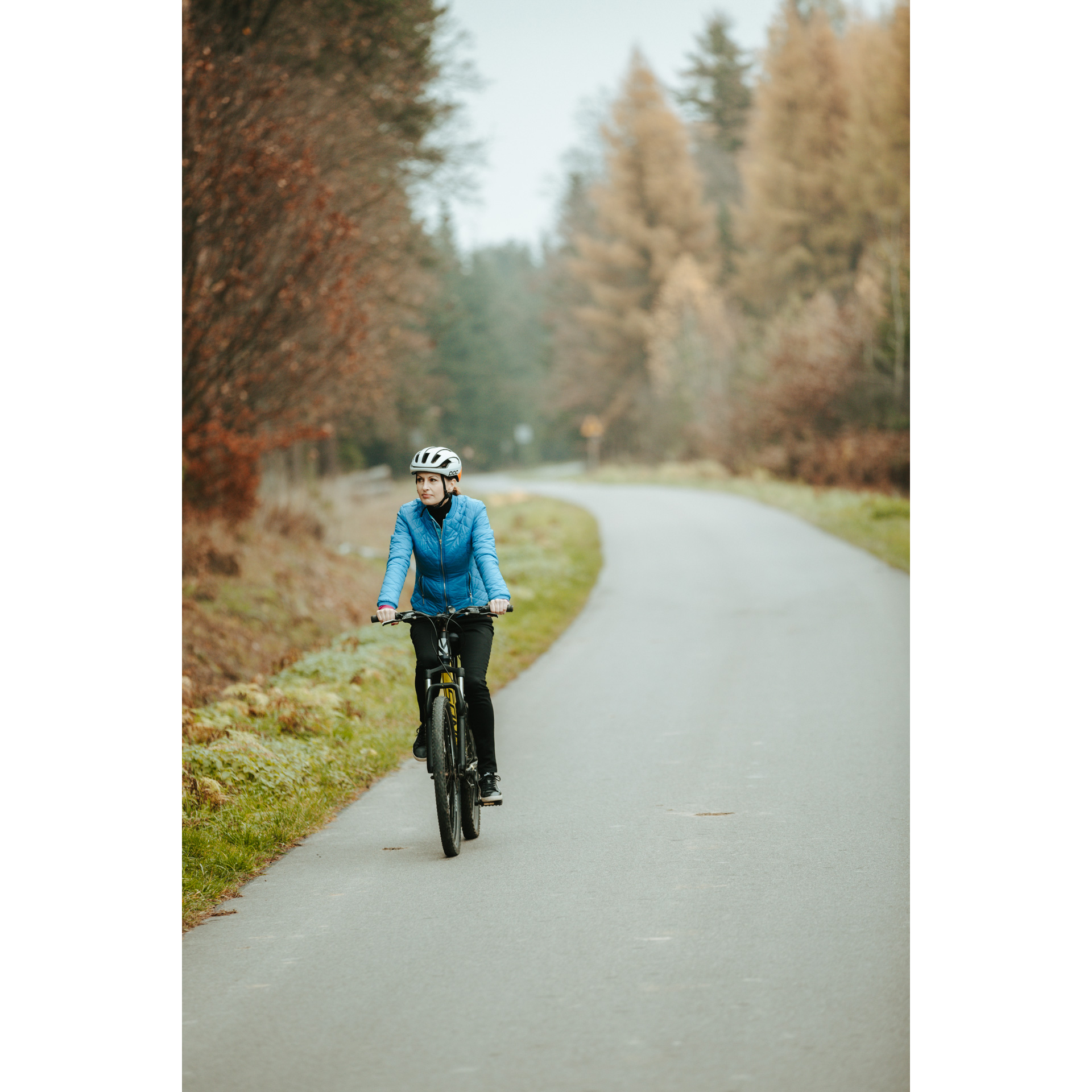 Rowerzystka w niebieskiej kurtce i białym kasku jadąca na rowerze asfaltową drogą, w tle kolorowe drzewa i krzewy