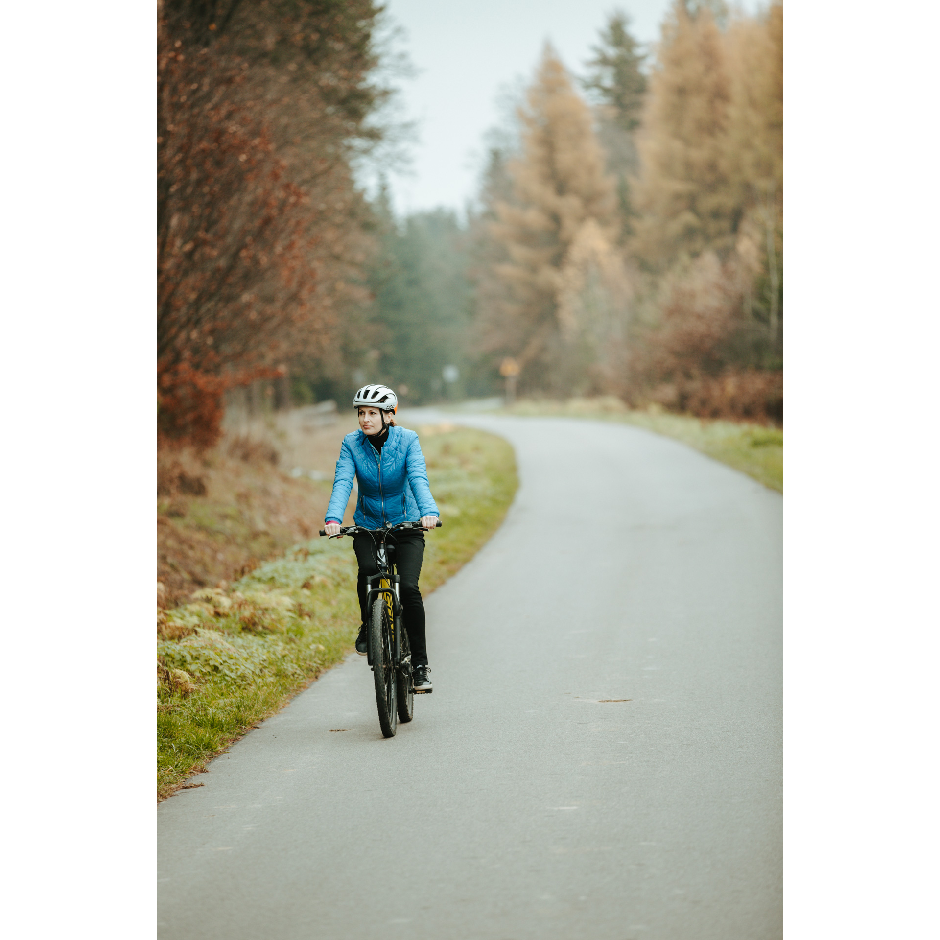 A cyclist in a blue jacket and a white helmet cycling along an asphalt road, colorful trees and bushes in the background