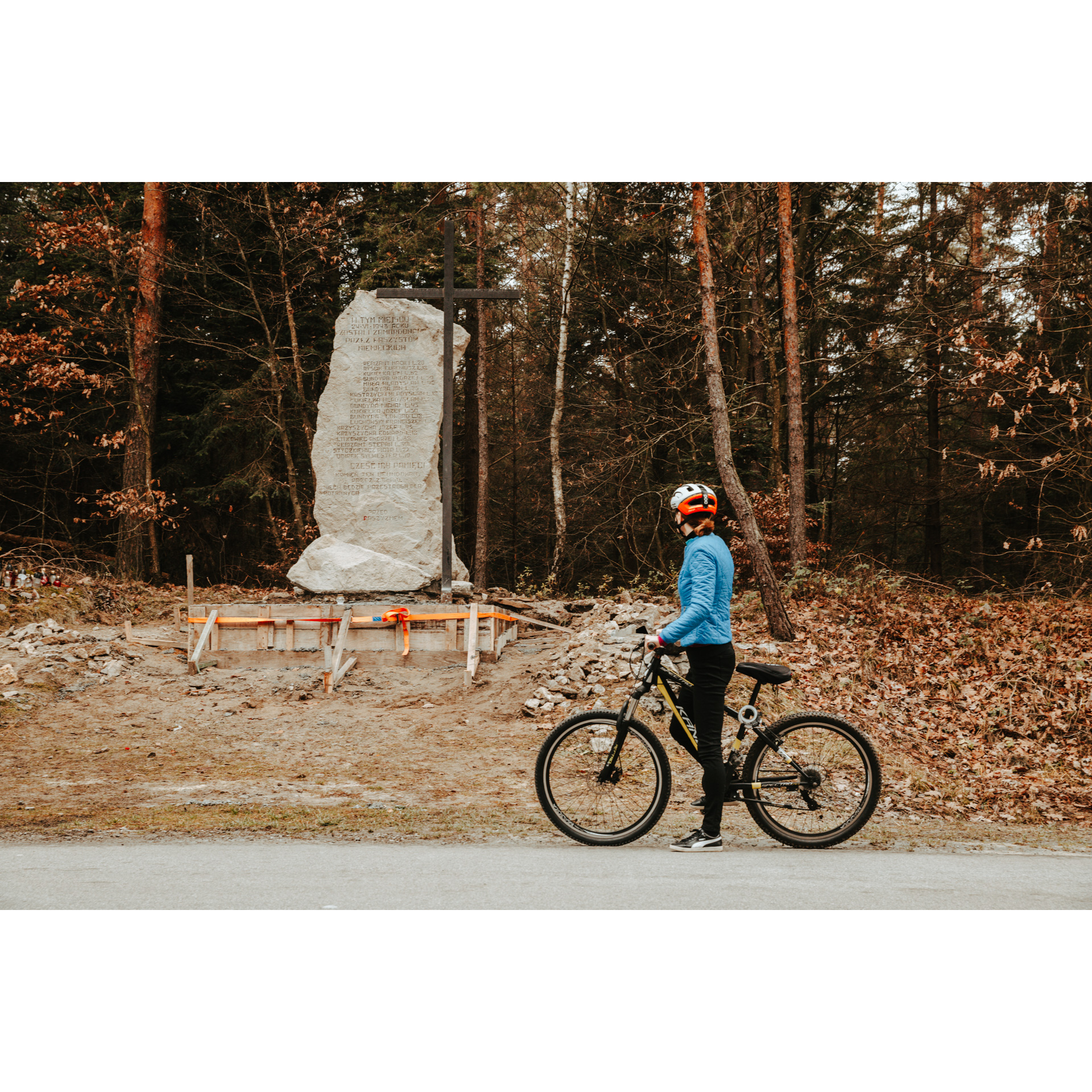 A cyclist in a blue jacket and a white helmet holding a bicycle and looking at a memorial stone standing by forest trees