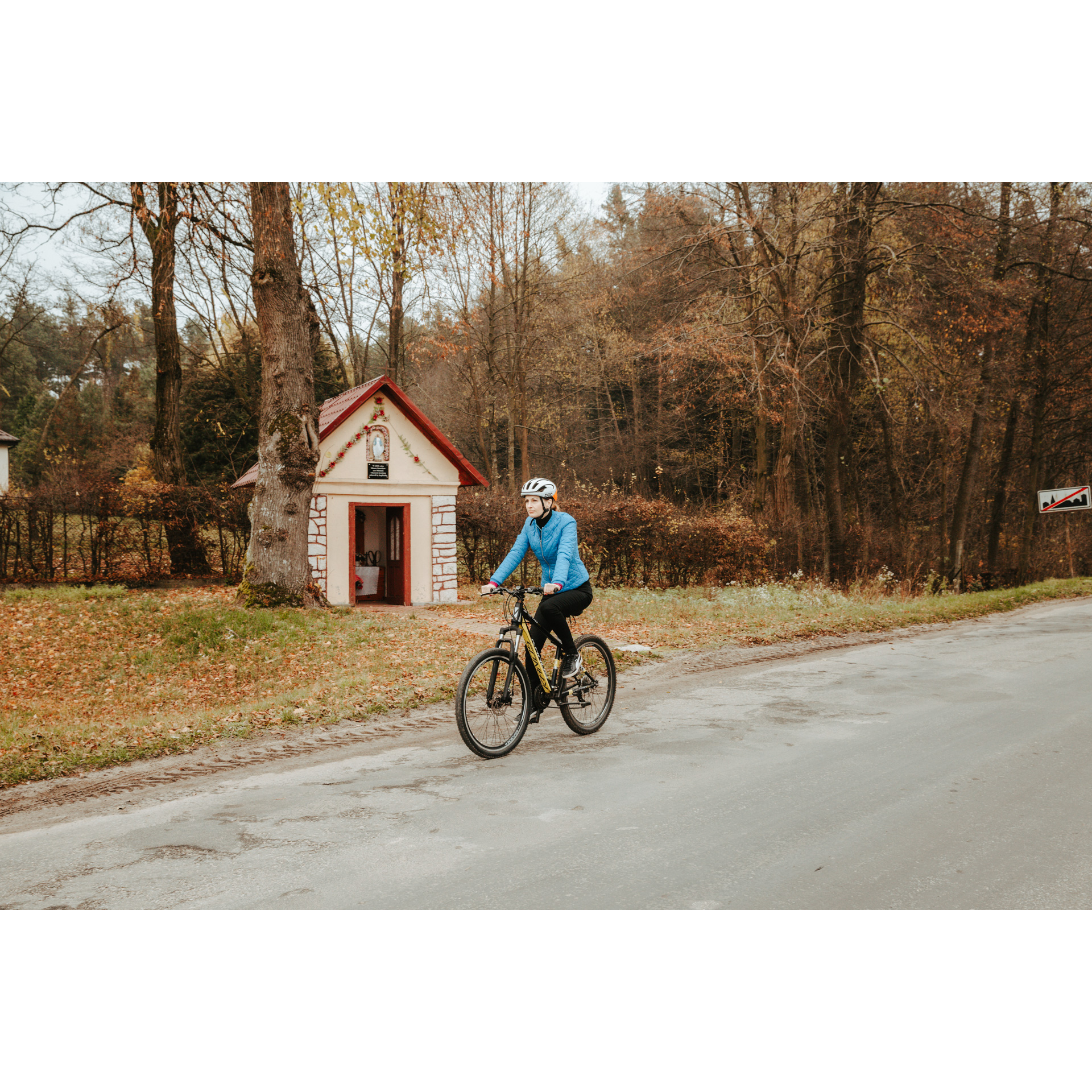 Rowerzystka w niebieskiej kurtce i białym kasku jadąca na rowerze asfaltową drogą, mijająca murowaną kapliczkę z czerwonym daszkiem