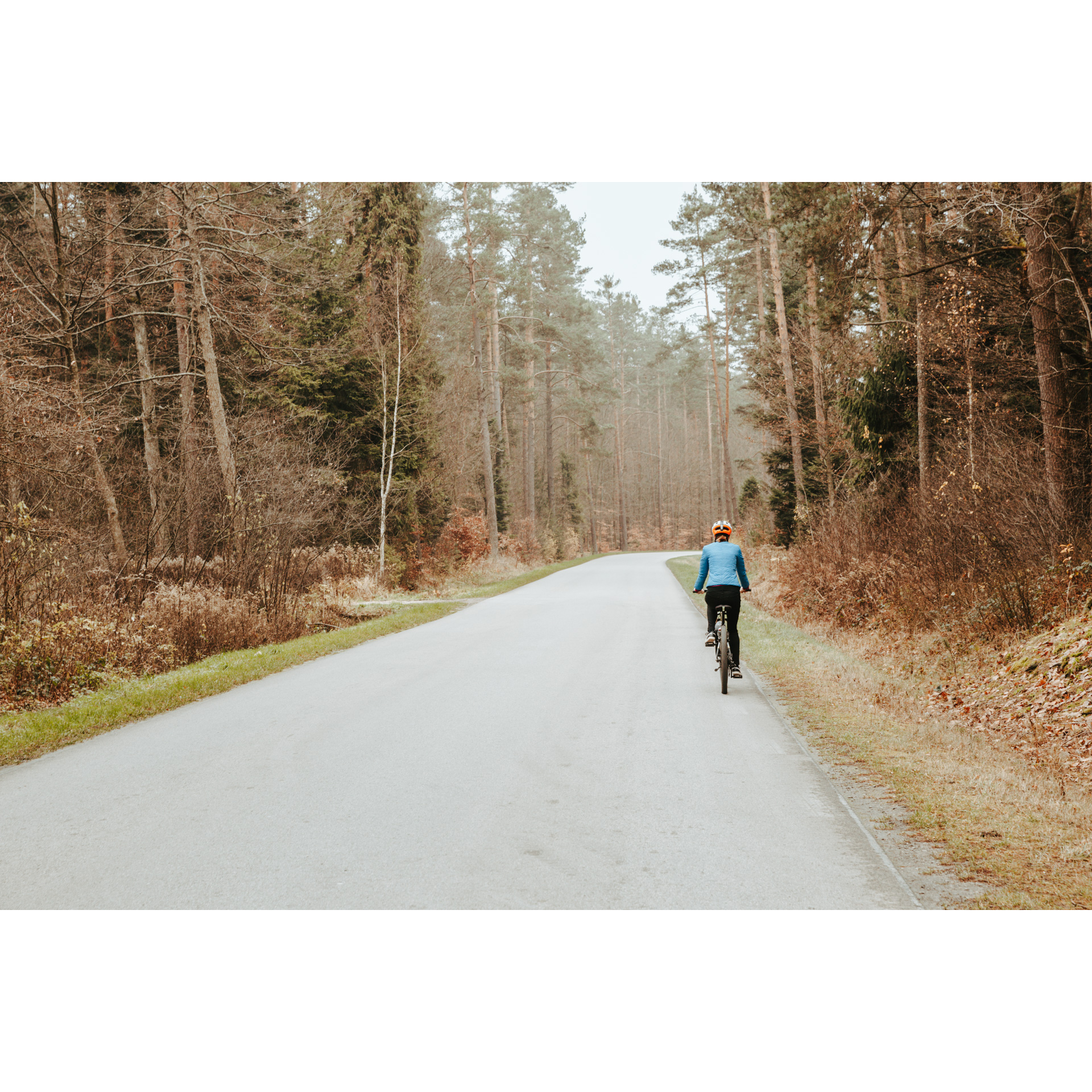 Rowerzystka w niebieskiej kurtce i białym kasku jadąca na rowerze asfaltową drogą wśród brązowo-złotych drzew i krzewów