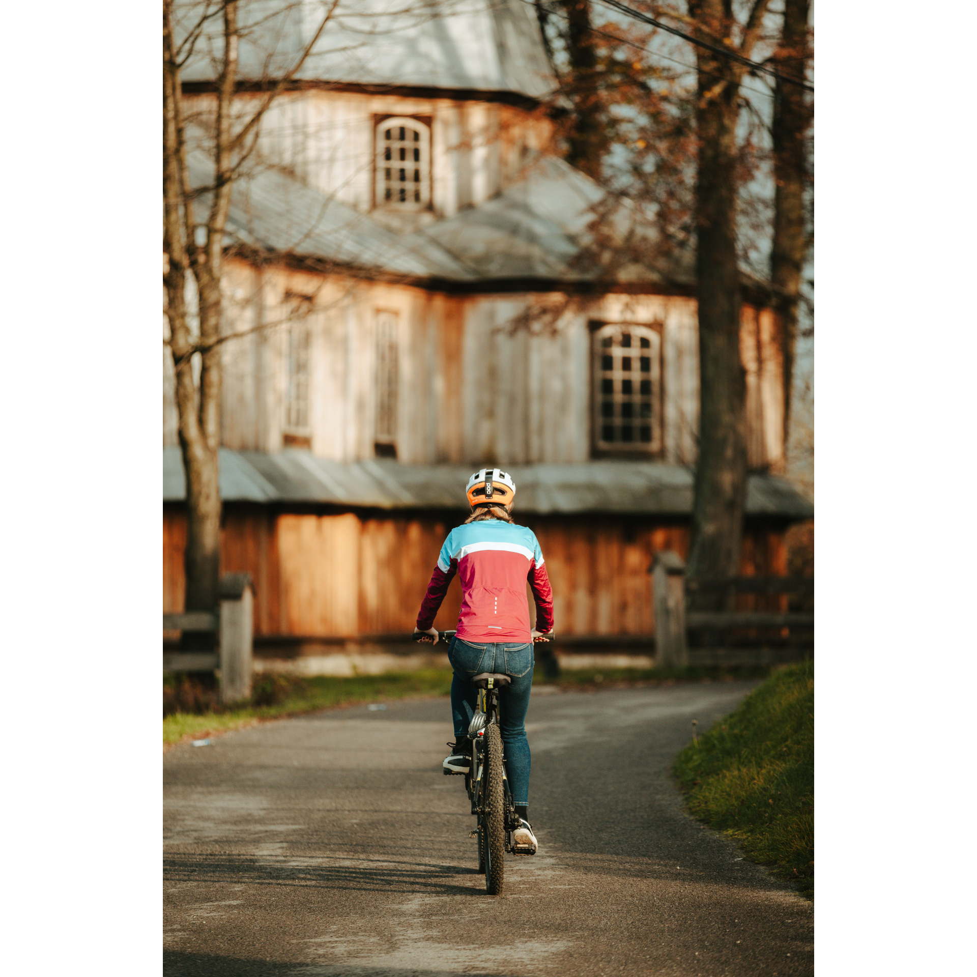 Rowerzystka w czerwono-niebieskiej kurtce i kasku jadąca rowerem po asfaltowej drodze, zmierzająca w stronę drewnianego kościoła