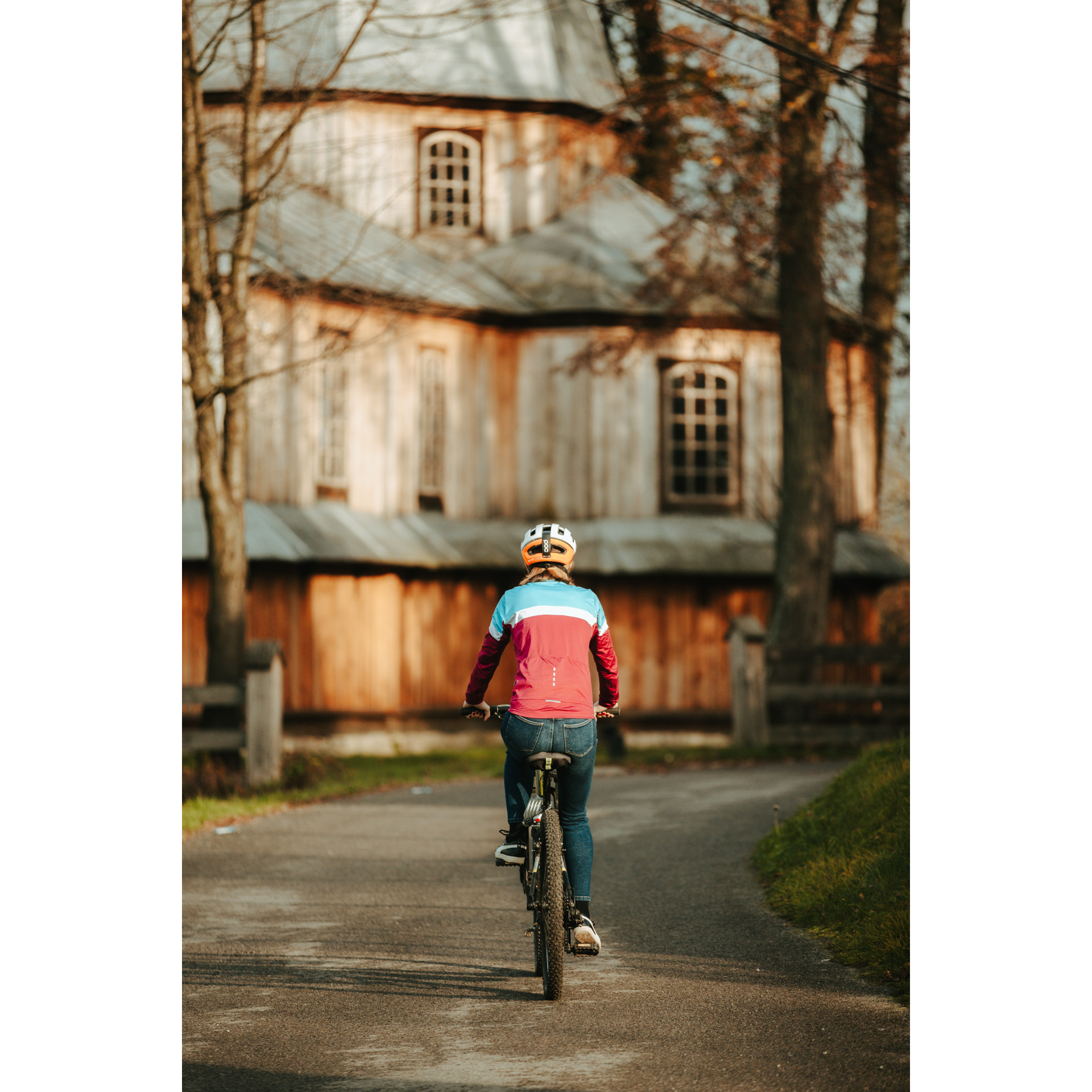 A cyclist in a red-blue jacket and helmet riding a bicycle on an asphalt road heading towards a wooden church