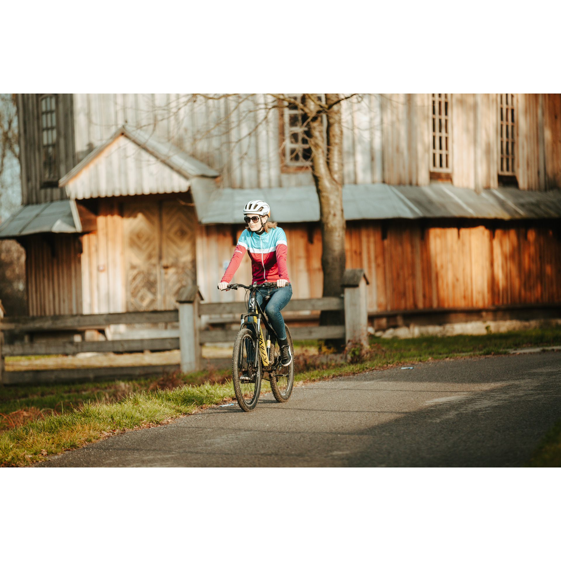Rowerzystka w czerwono-niebieskiej kurtce i kasku jadąca rowerem po asfaltowej drodze, mijająca drewniany budynek kościoła