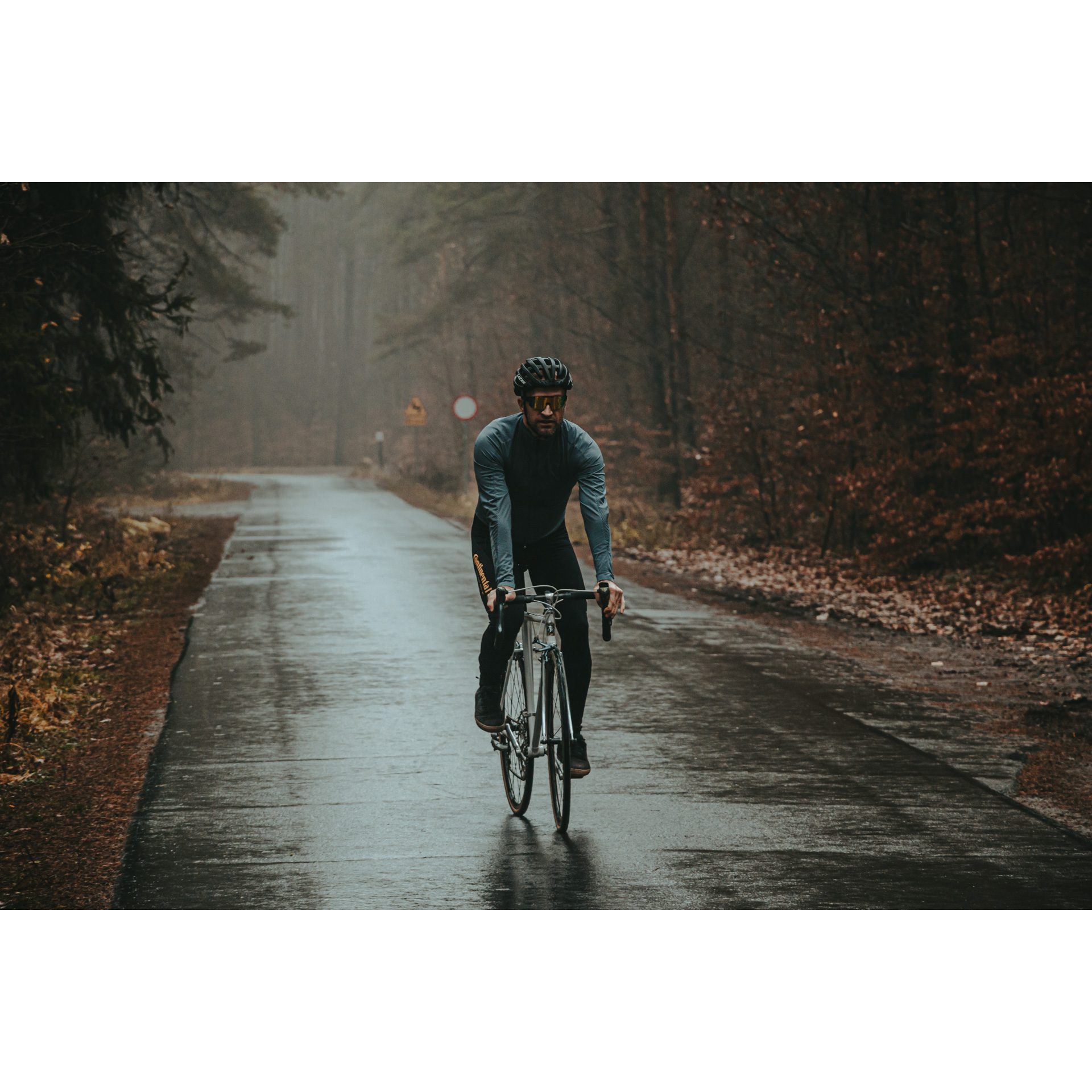 A cyclist in a gray and black outfit and a helmet riding a bicycle on a forest, wet, asphalt road