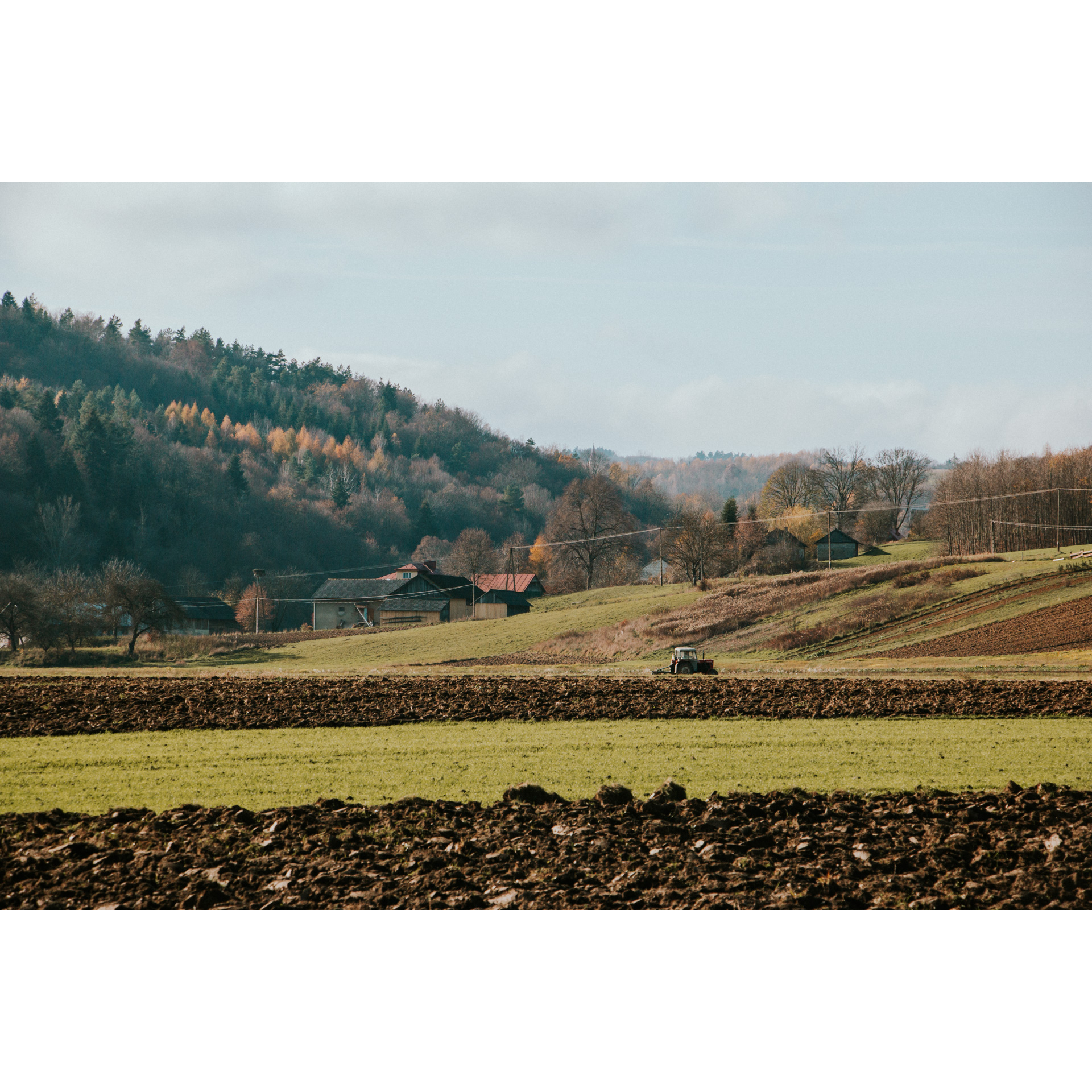 View of fields and meadows, in the background a moving tractor, outbuildings and a hill covered with trees and a bright sky