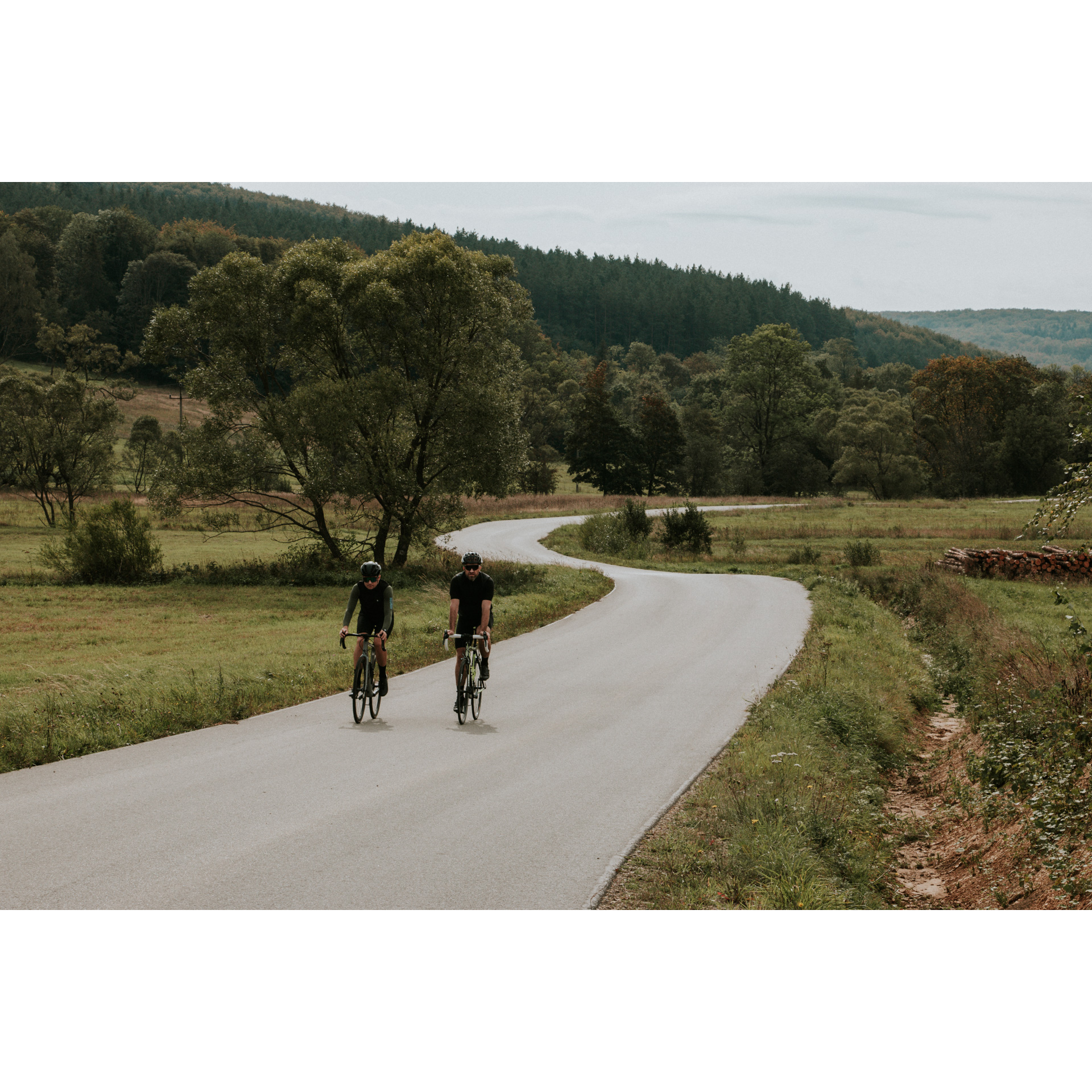 Dwóch kolarzy w czarnych strojach i kaskach jadących na rowerach asfaltową krętą drogą wśród zielonych łąk, drzew i wzgórz