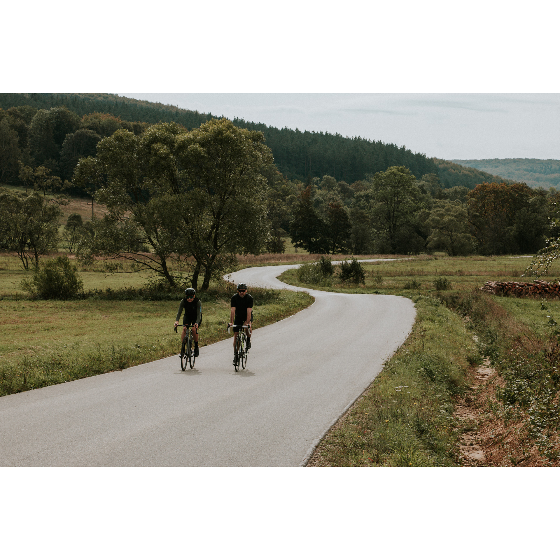 Two cyclists in black clothes and helmets riding bicycles along an asphalt winding road among green meadows, trees and hills