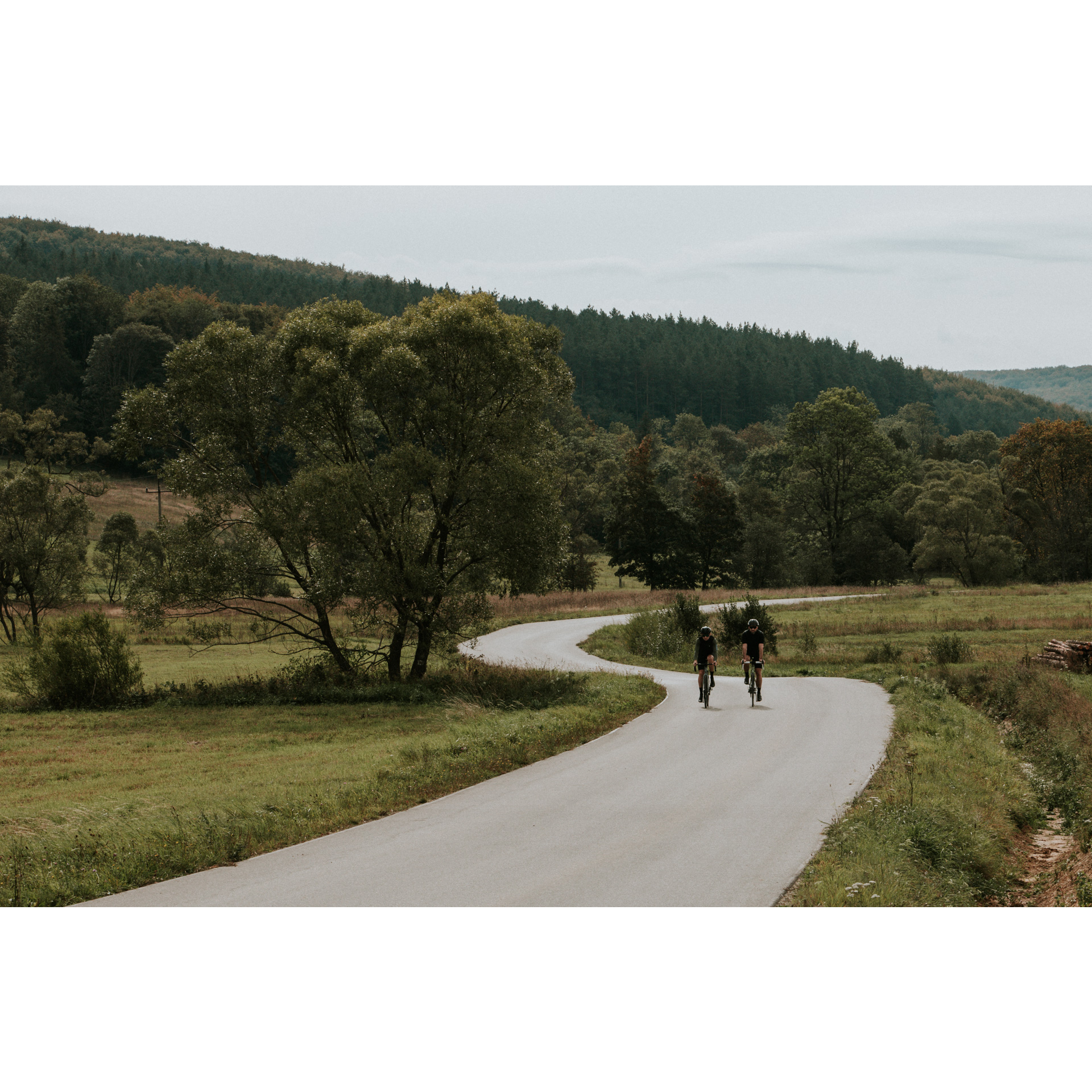Dwóch kolarzy w czarnych strojach i kaskach jadących na rowerach asfaltową krętą drogą wśród zielonych łąk, drzew i wzgórz
