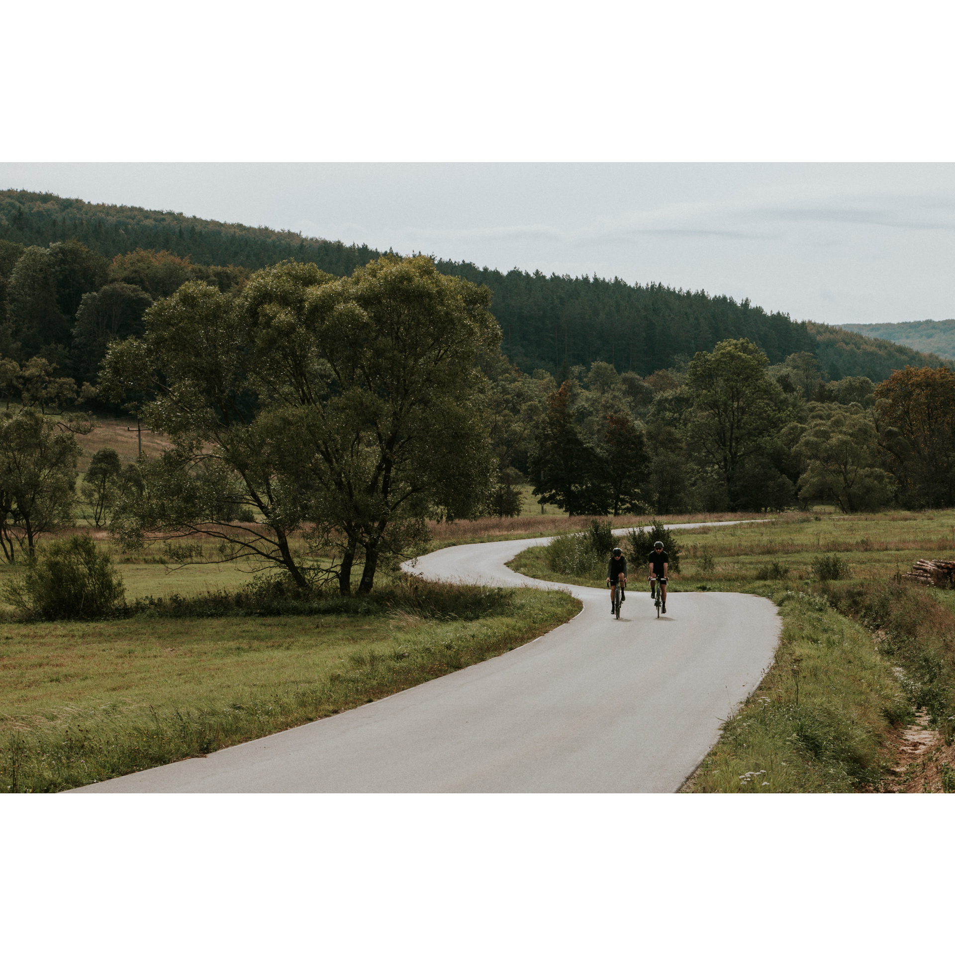 Two cyclists in black clothes and helmets riding bicycles along an asphalt winding road among green meadows, trees and hills